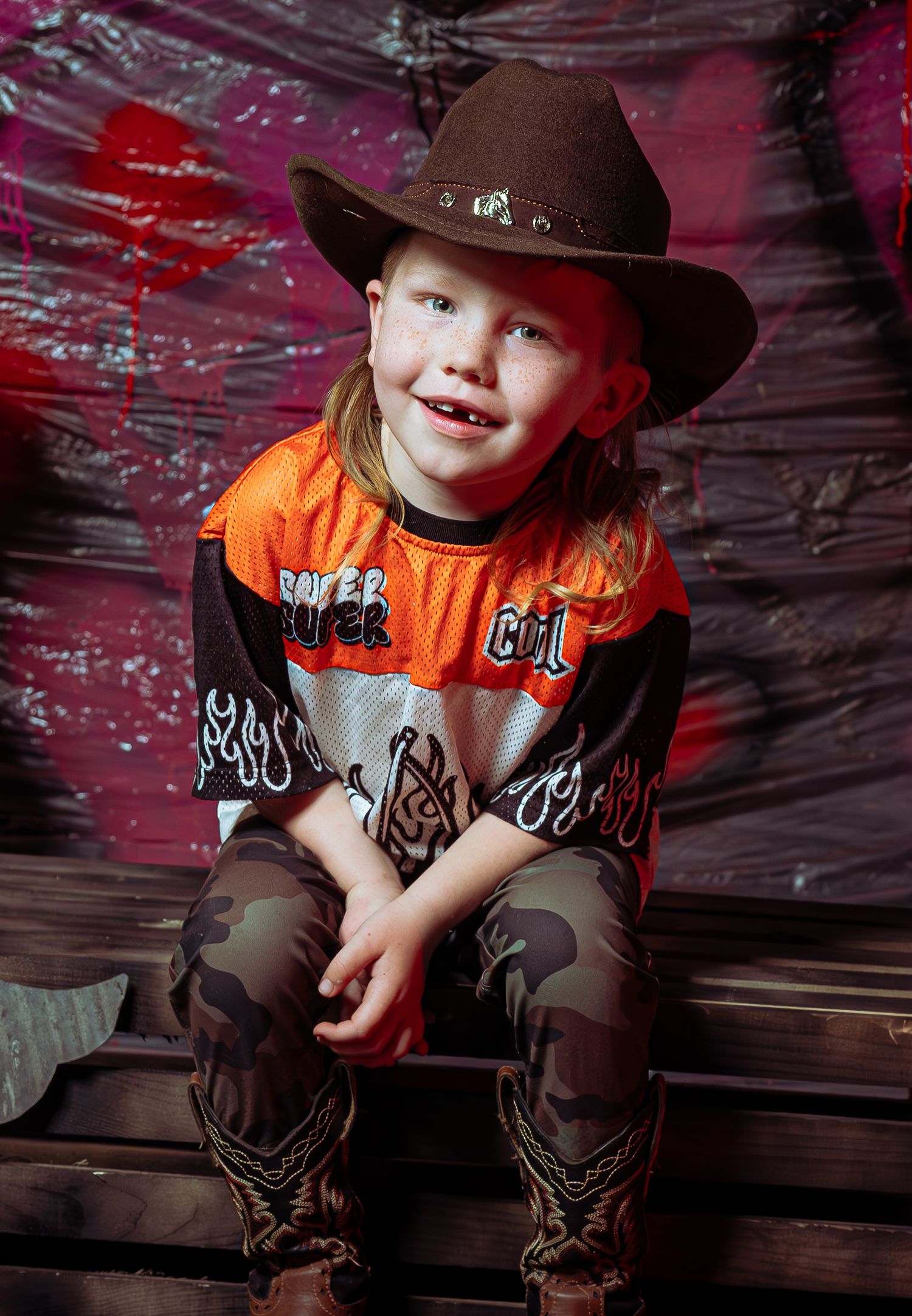 A young child wearing a brown cowboy hat, orange shirt and brown pants sits against a dark rustic background.