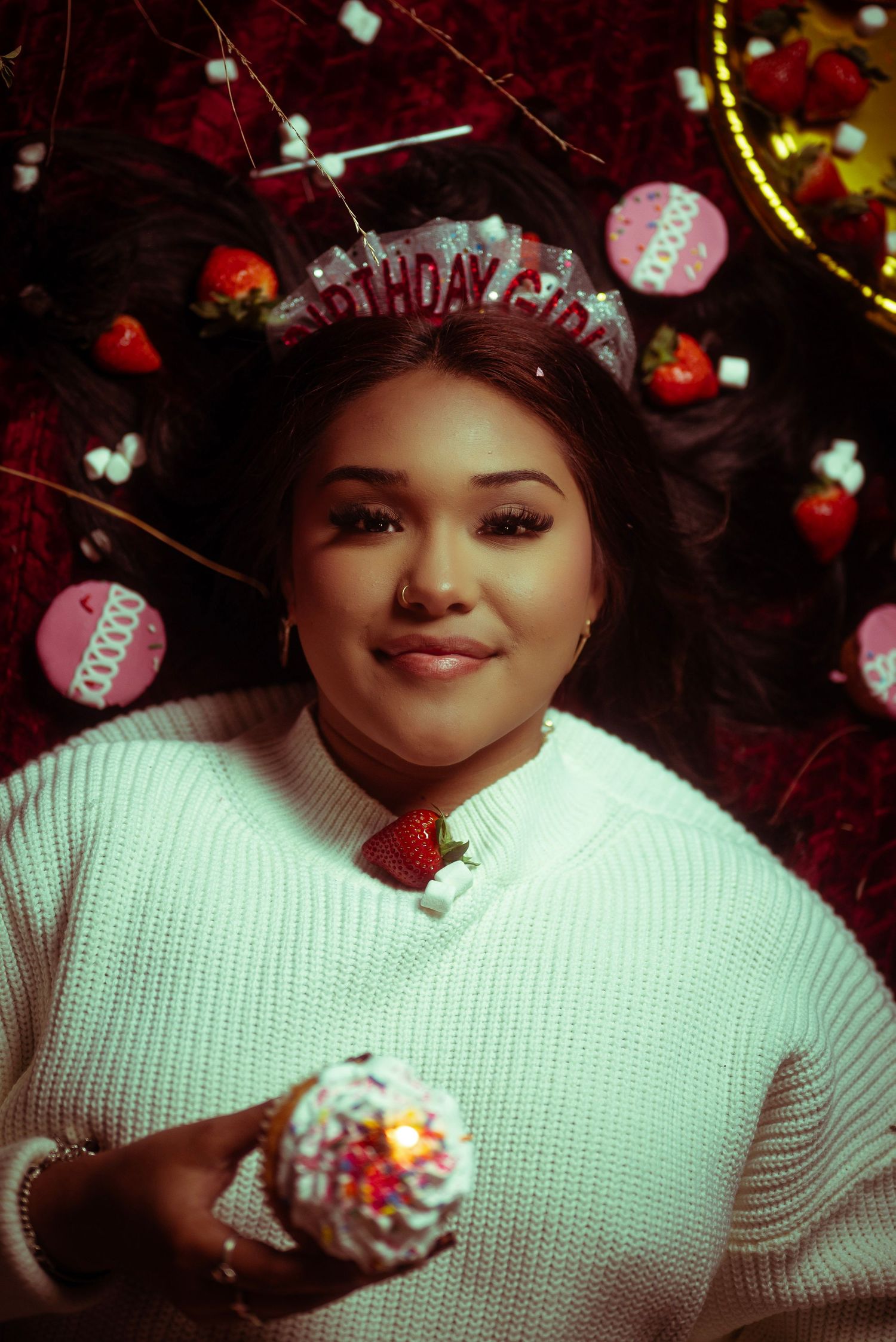 A person in a white sweater poses with festive holiday decorations and birthday elements in a dark setting.