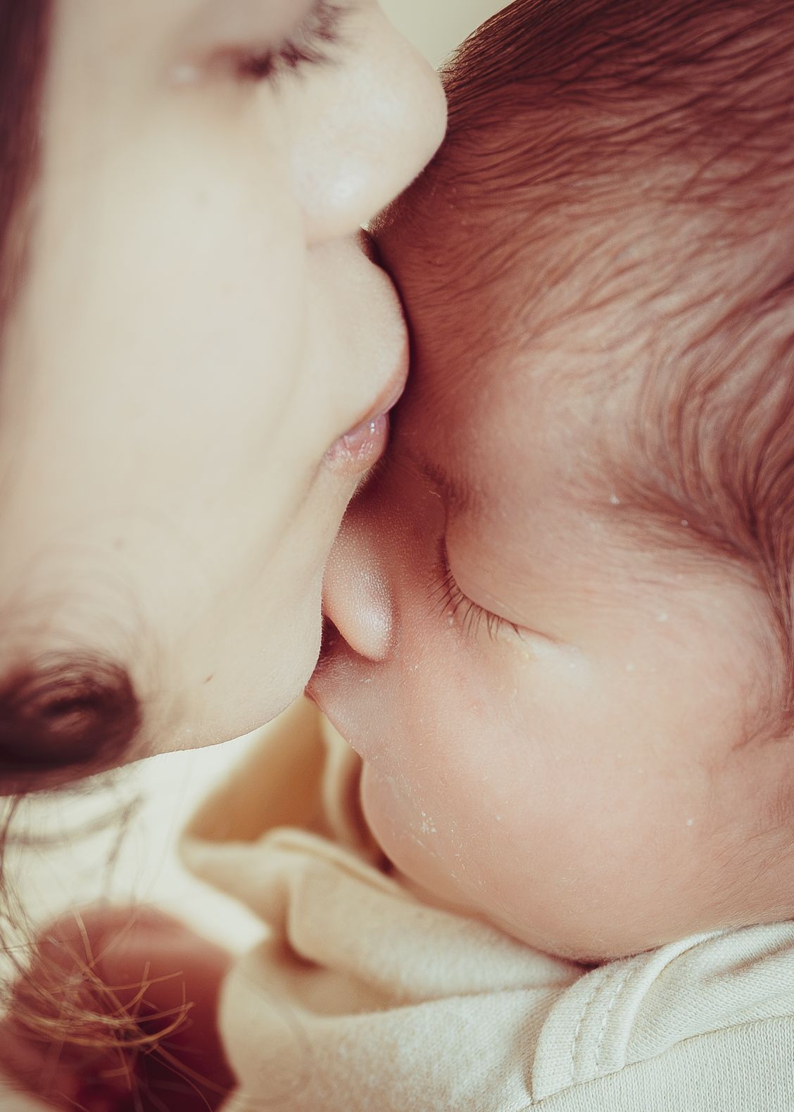 A tender close-up of a mother gently kissing her sleeping newborn baby's head.