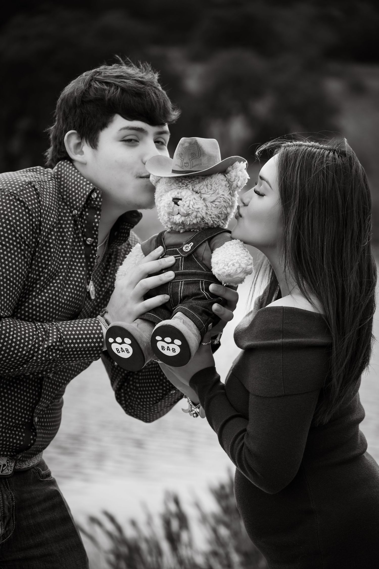 Black and white photo of a couple kissing a teddy bear wearing a cowboy hat outdoors.