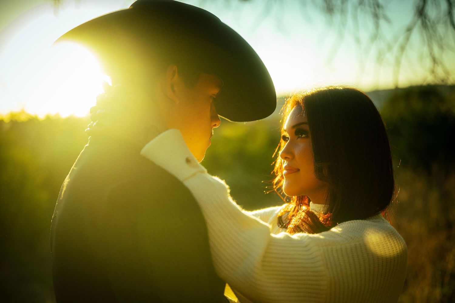 Couple sharing a romantic moment outdoors, bathed in golden sunset light, man wearing cowboy hat, woman in white sweater.