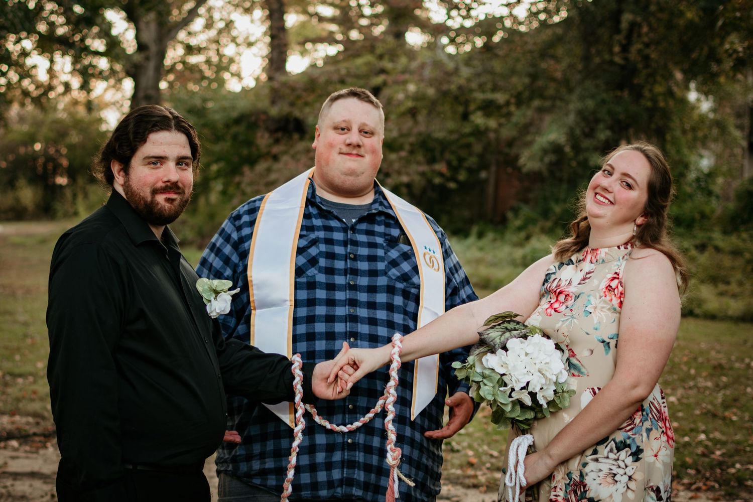 Wedding- The bride, groom, and officiant look happily at the camera. Bride and groom are holding hands.