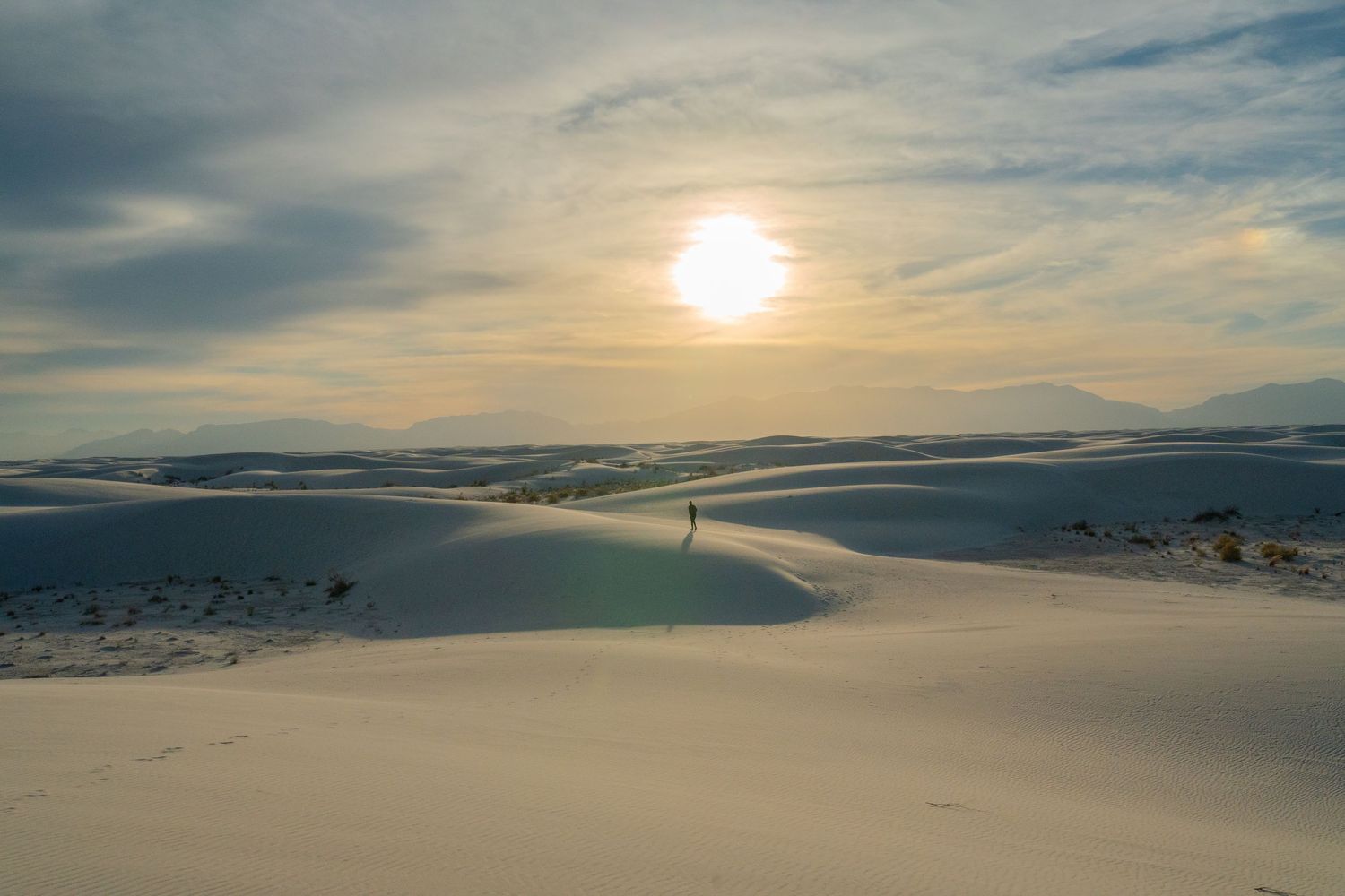 Image of a bright sun as it sets, hidden behind thin clouds. Huge sand dunes are going as far as the eye can see, a person in the distance.