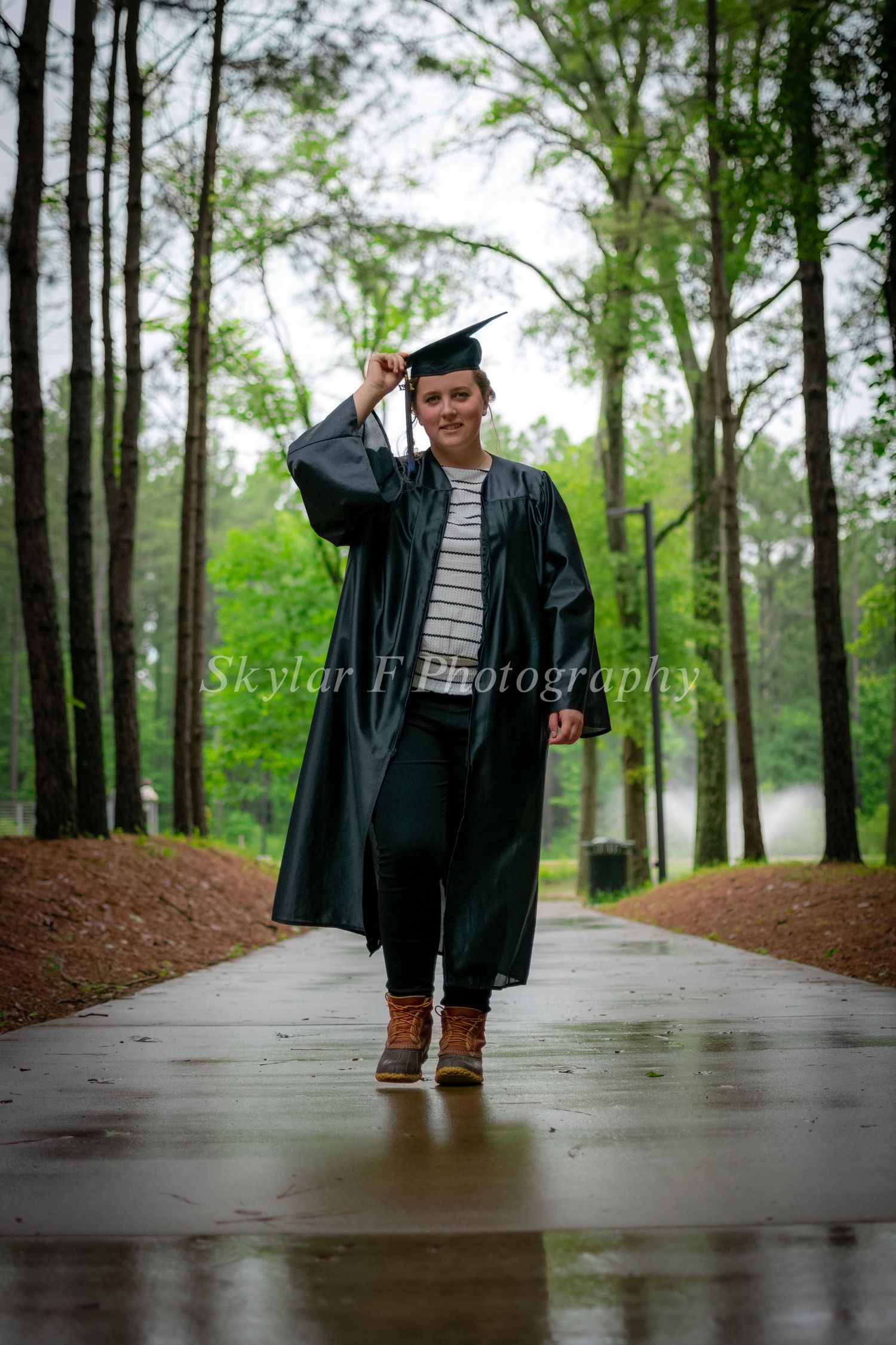 A girl in a cap and gown holds her cap and walks toward the camera on a rainy sidewalk surrounded by bright green leaves.