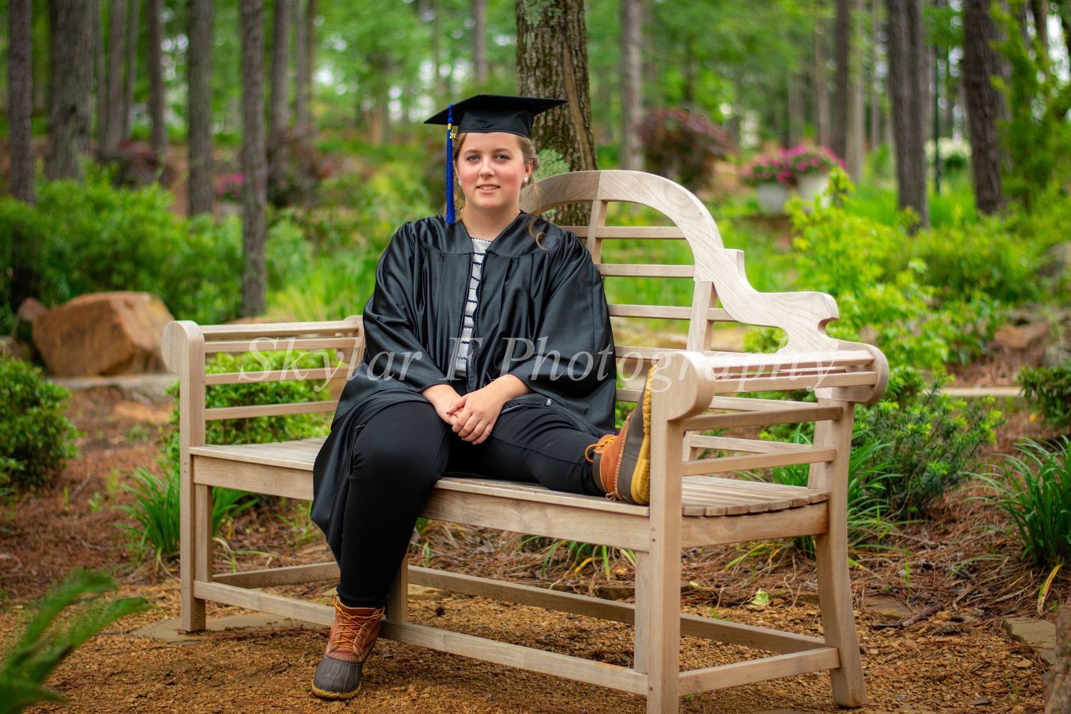 Surrounded by bright green foilage, a girl wearing a black cap and gown sits with one leg on a bench and on leg on the floor looking serene.