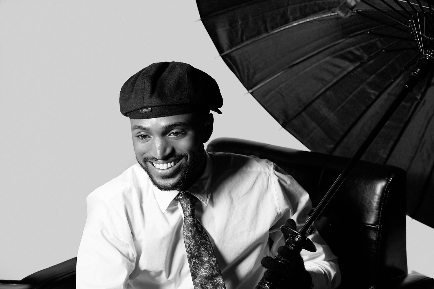 Black and white portrait of someone in a flat cap and tie holding an umbrella against a studio backdrop.