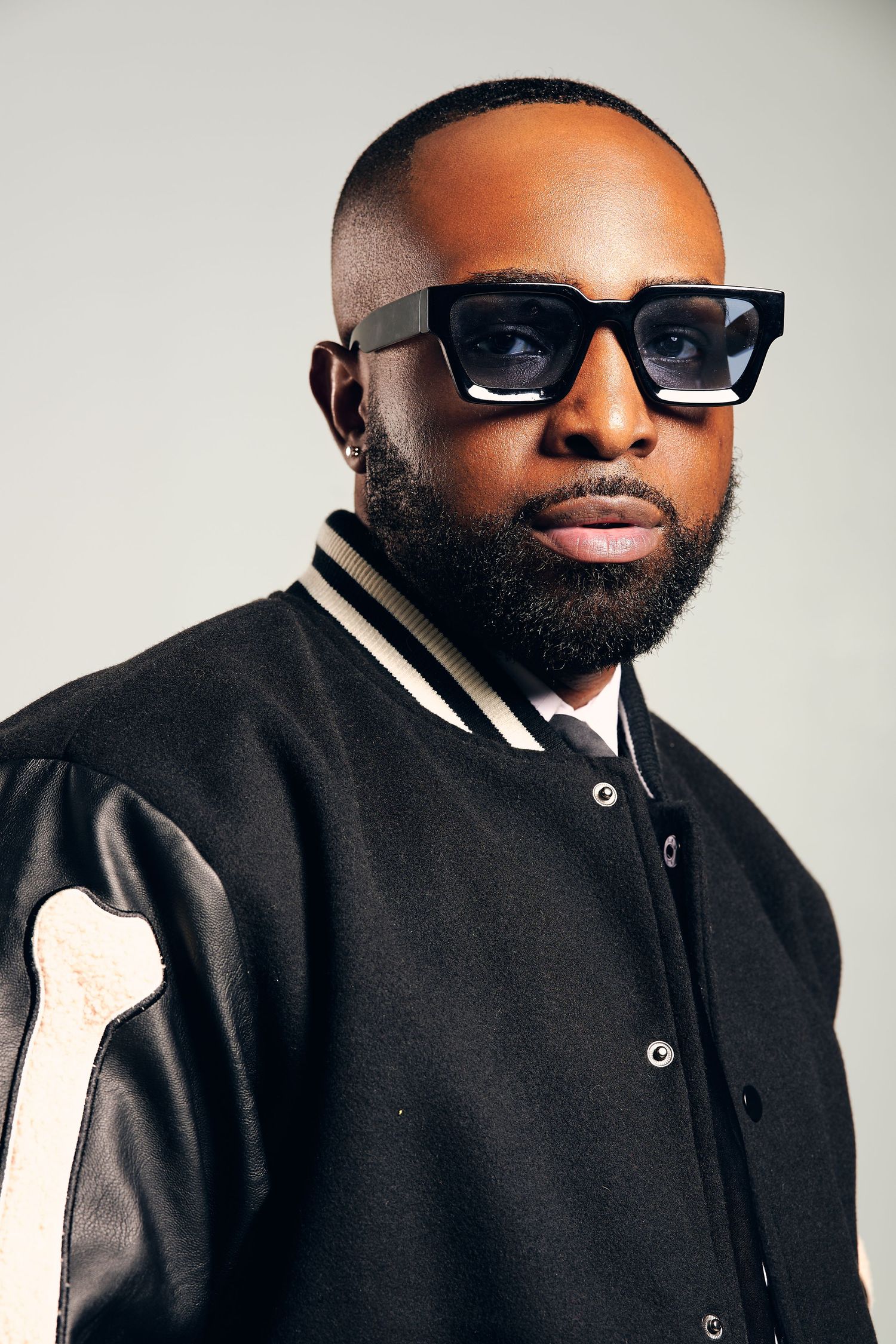 Fashion portrait series featuring black varsity jacket and angular sunglasses against light background.