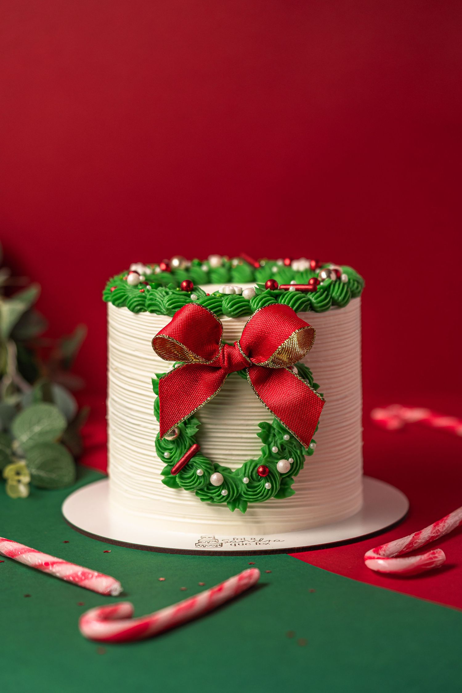 White Christmas cake decorated with green wreath and red bow against red background.