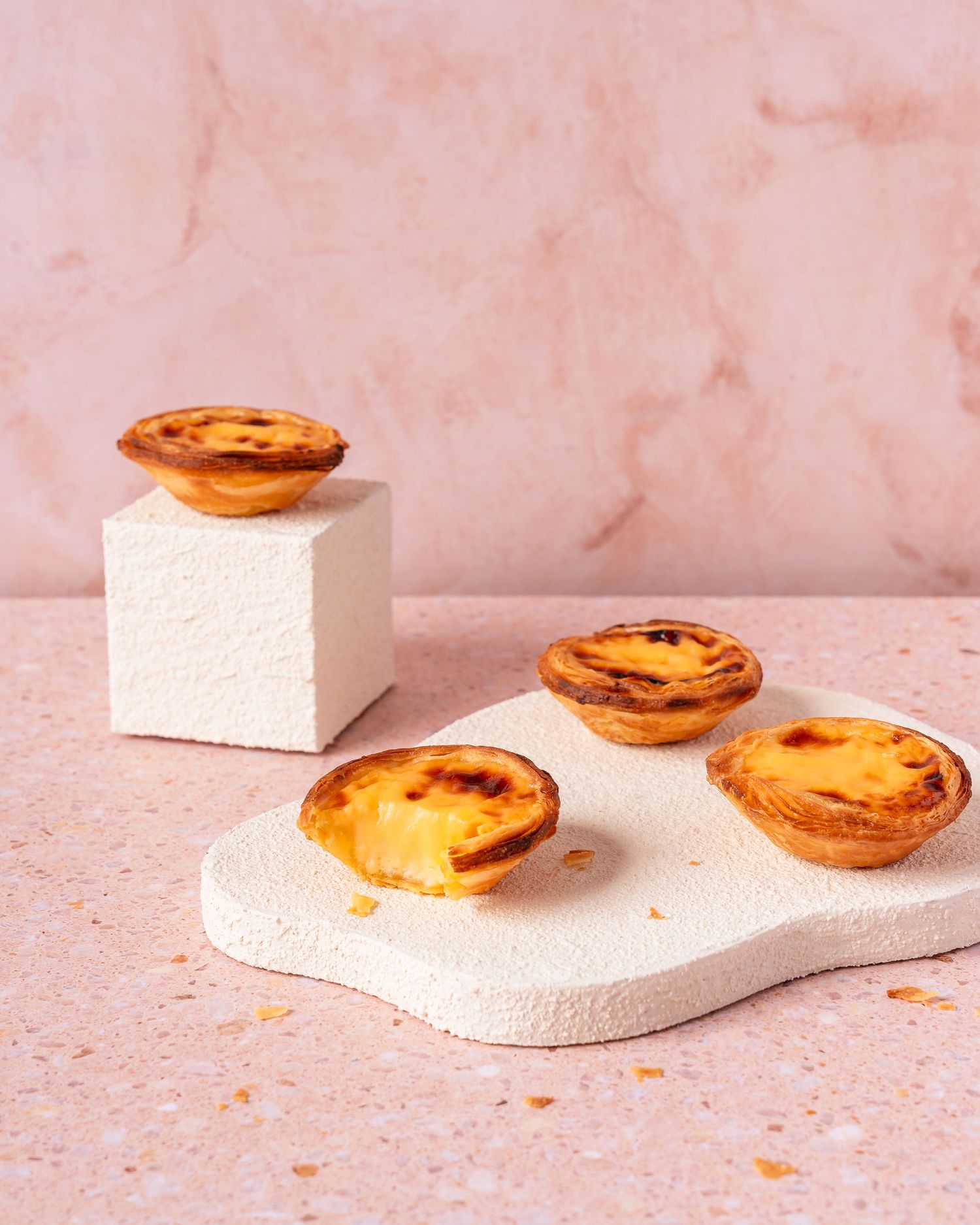 Portuguese egg tarts arranged on white geometric platforms against pink background.