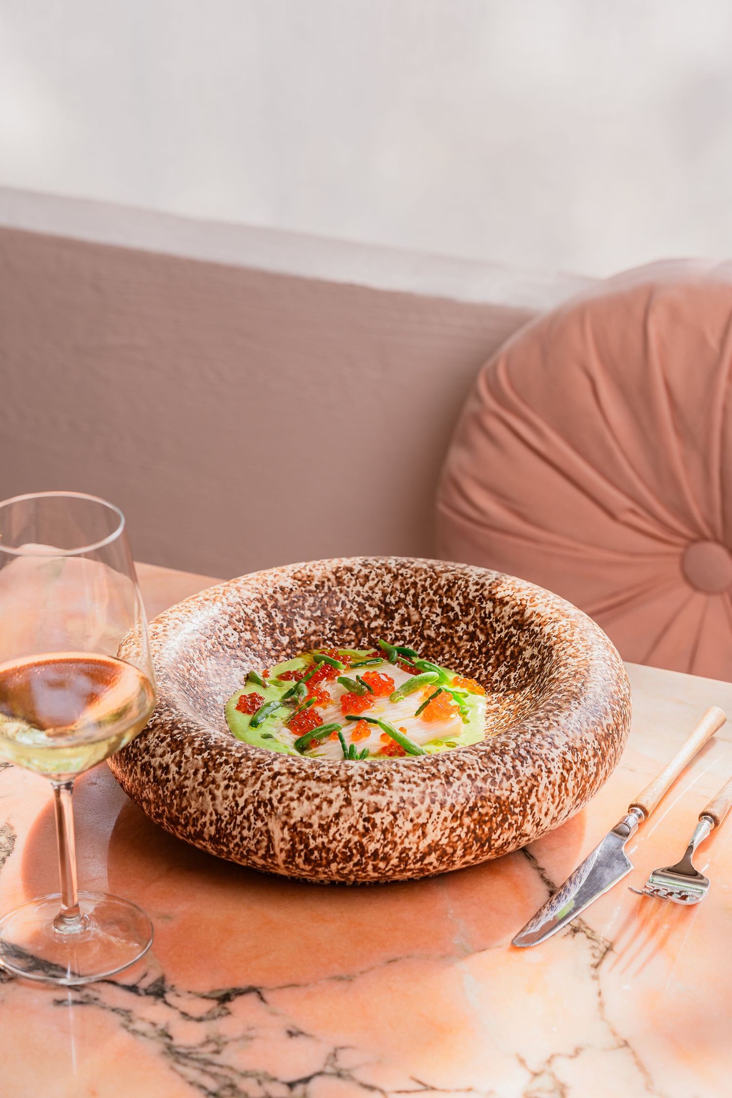 Elegant plated dish in textured bowl with wine glass on coral-colored surface.