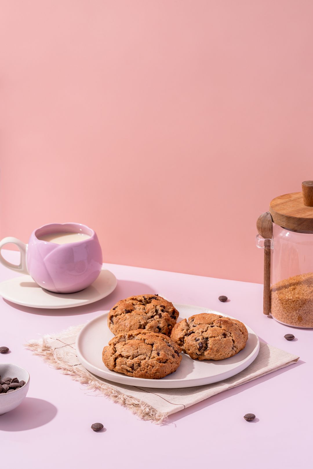 Chocolate chip cookies arranged on white plates against a pink background with scattered crumbs and oats.