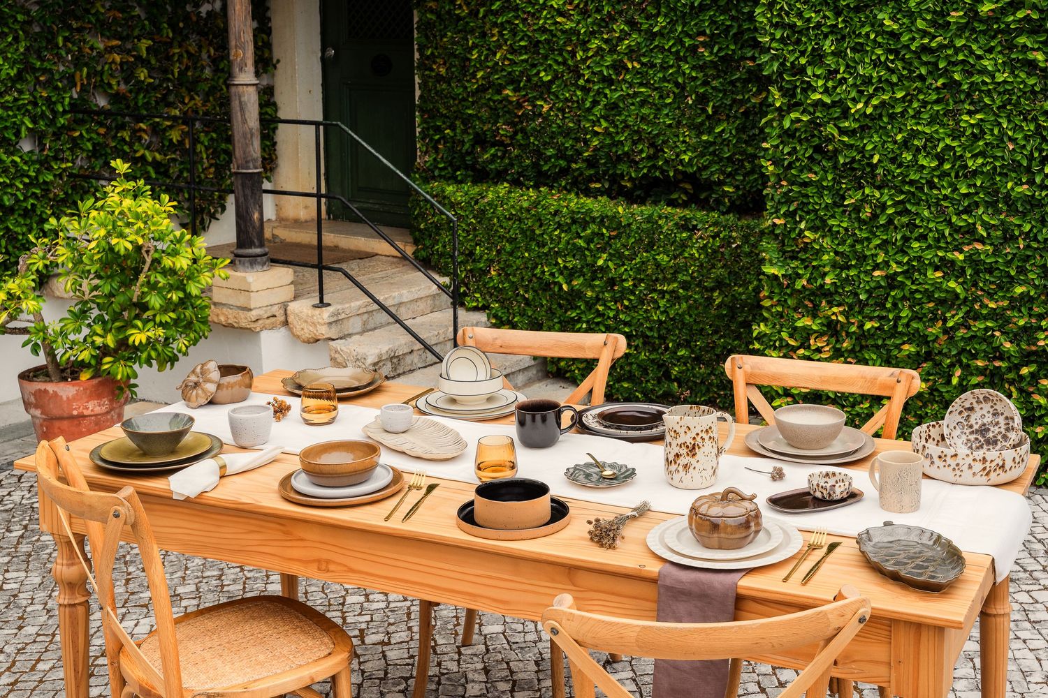 Long outdoor dining table set up in garden with green hedge backdrop.