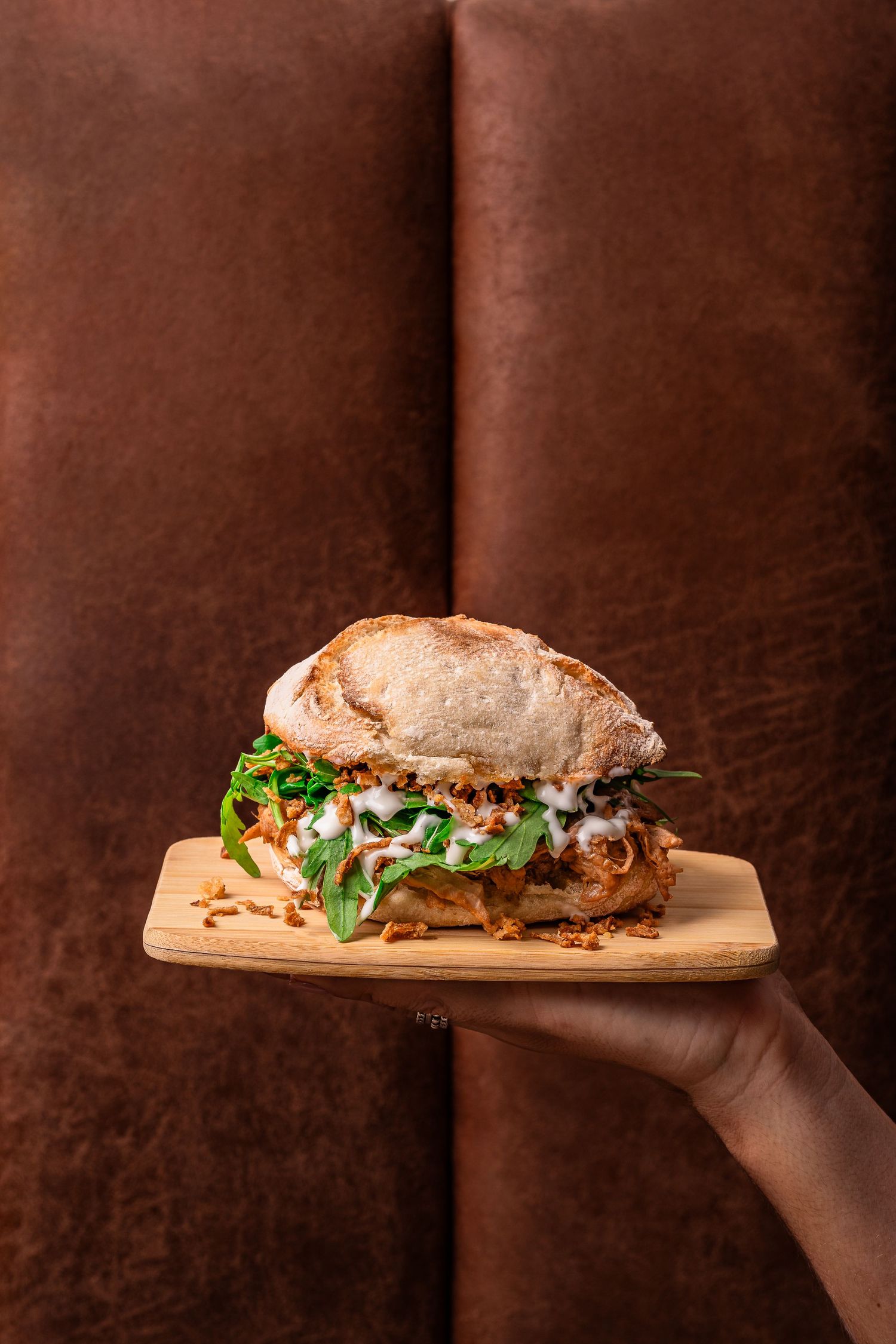 Long rustic sandwich on wooden board being held against dark background.