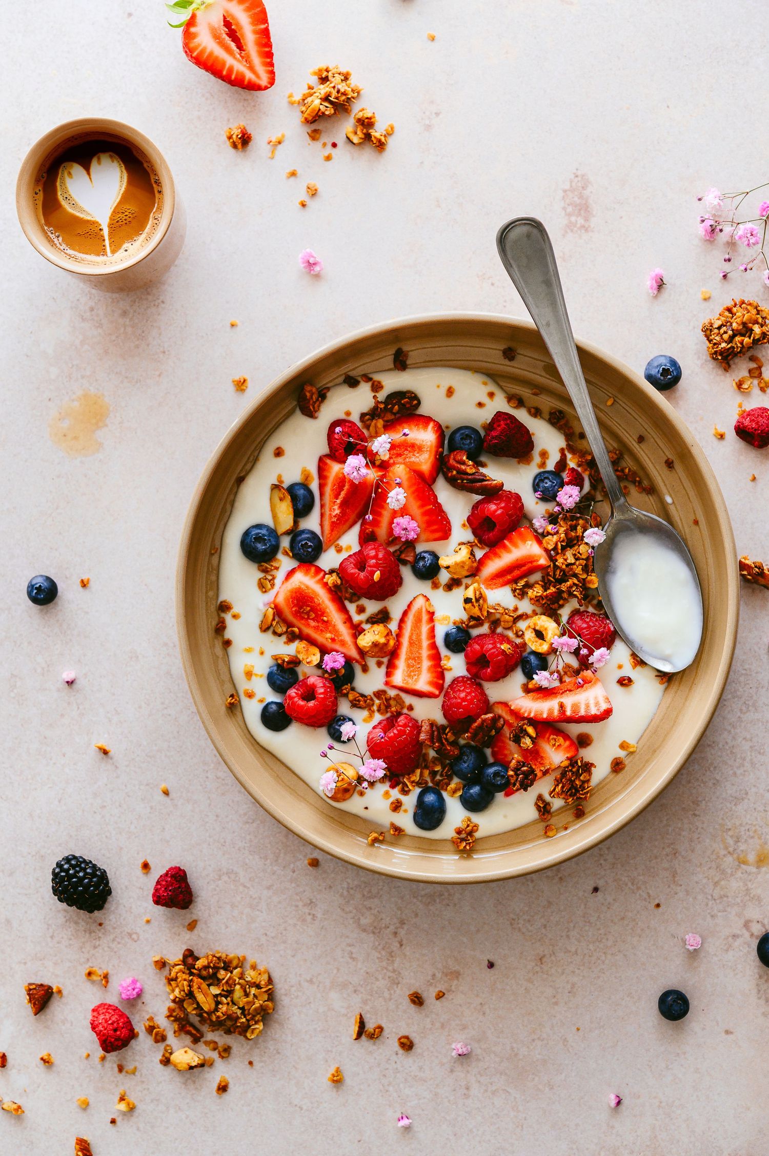 Breakfast bowl with yogurt, fresh berries, granola and latte art coffee on white surface.