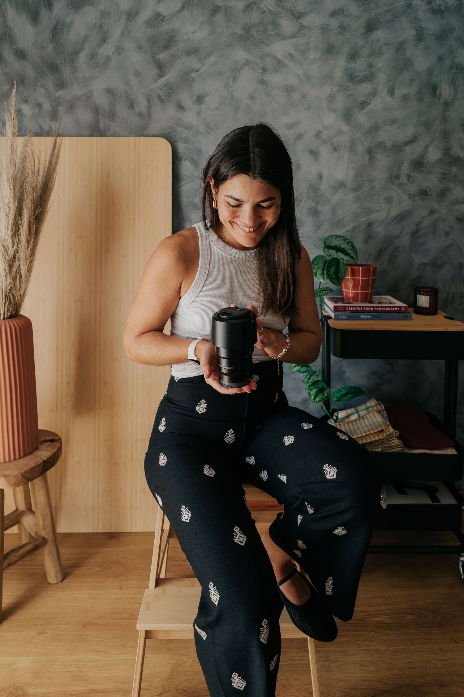 Person in white tank top and patterned pants holding a lens in a studio setting.