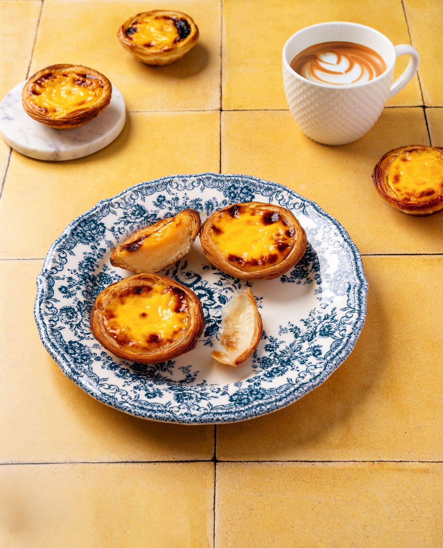 Portuguese egg tarts served on blue and white china plates with coffee on yellow tile surface.