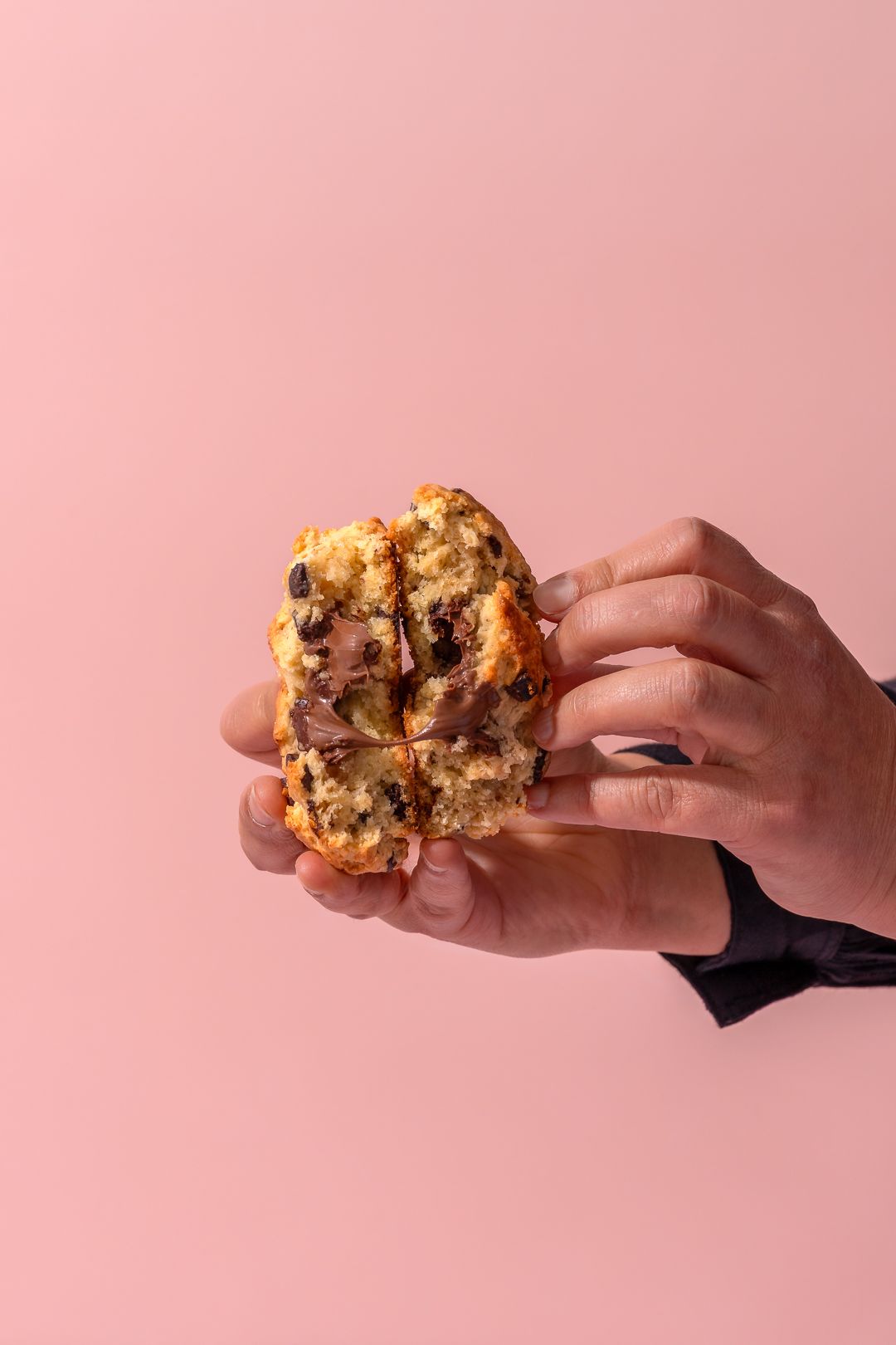 Hands holding a freshly baked chocolate chip cookie against pink background.