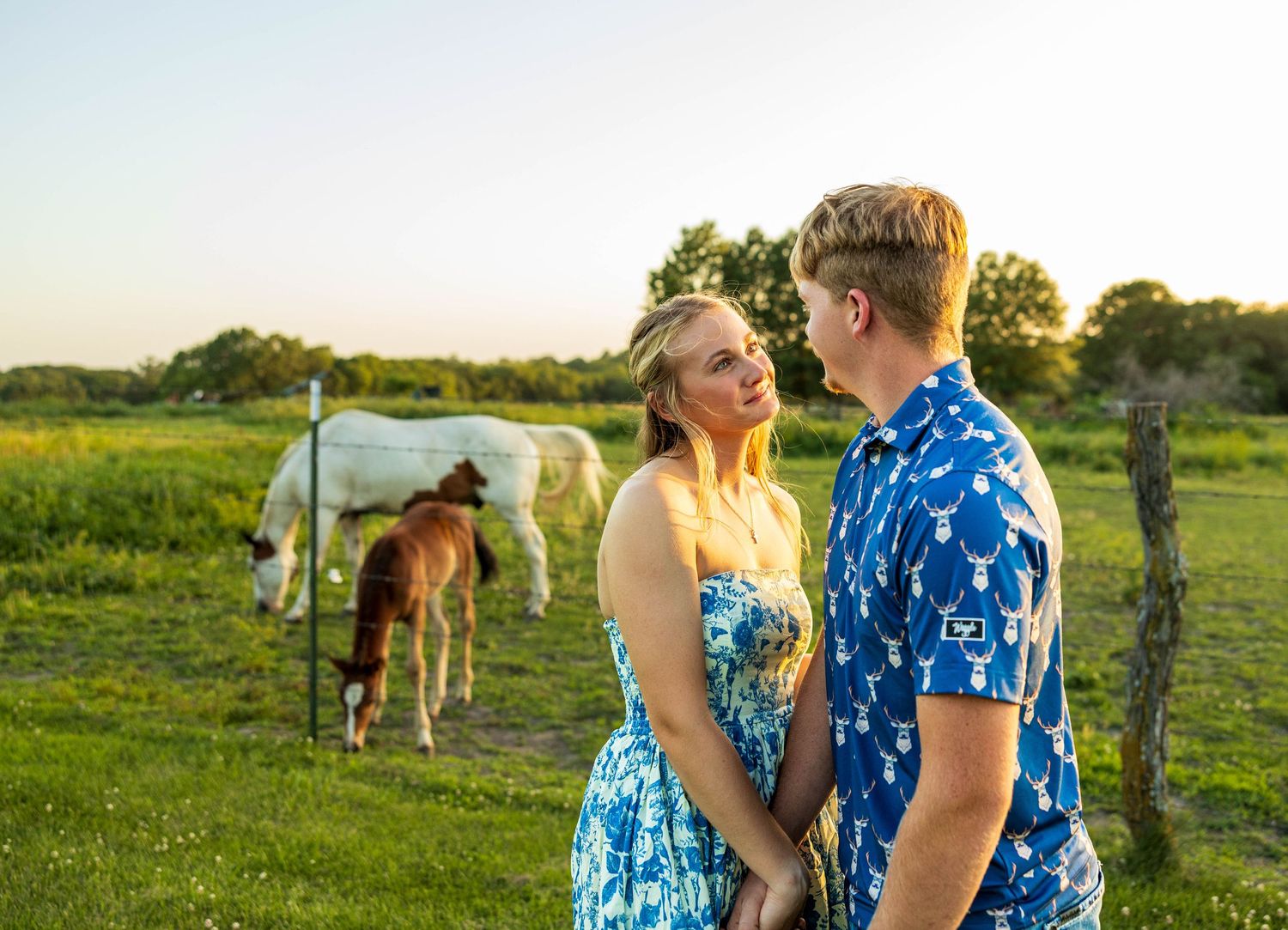 A couple looking at each other, holding hands in front of horses in a pasture.