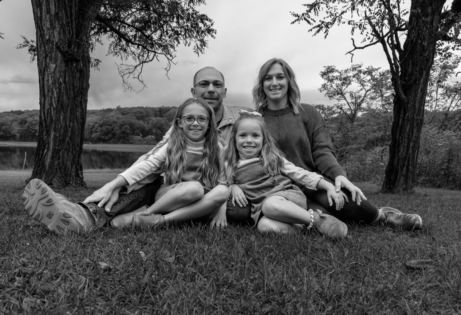 A young family sitting on the ground, with two little girls in a treed area.