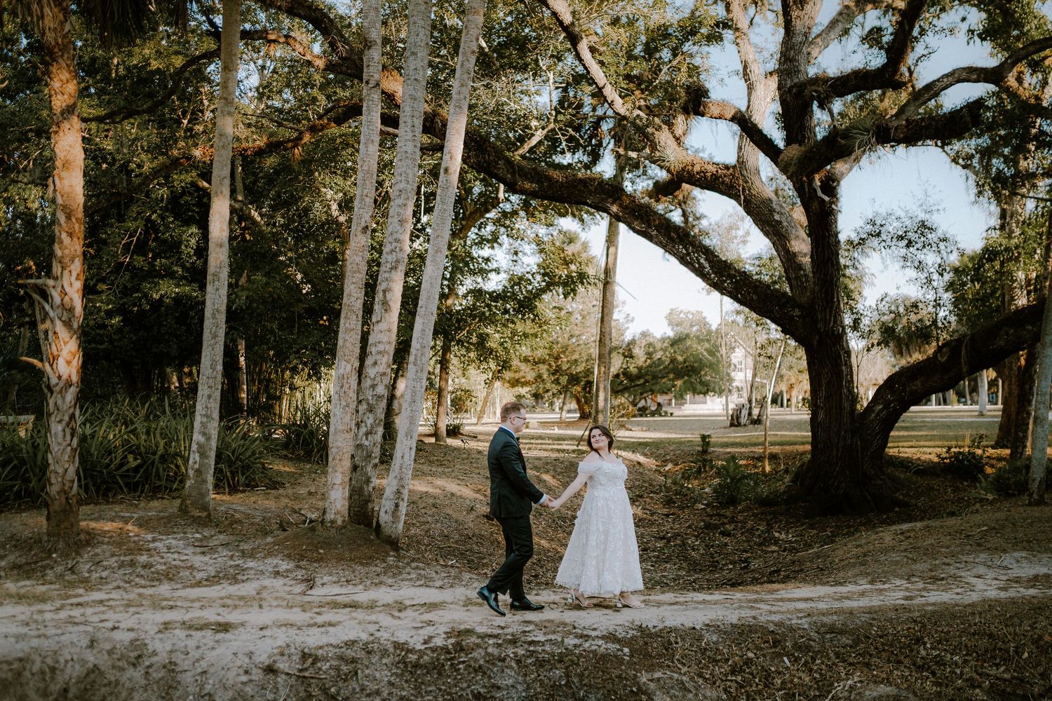 Bride and groom hold hands walking through a scenic tree-lined sandy path during their outdoor wedding celebration.