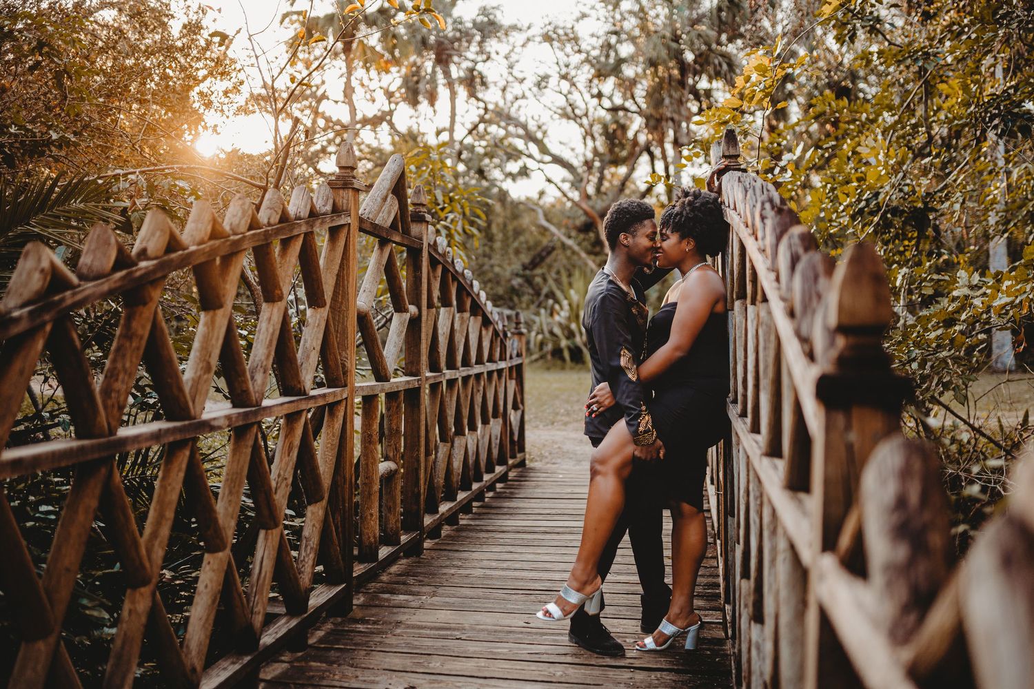 Couple sharing a romantic kiss on a wooden bridge surrounded by lush greenery in golden sunset light.