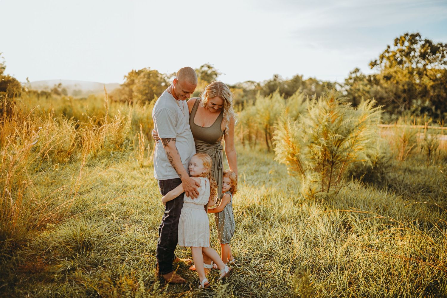A golden hour outdoor family portrait in tall grass with warm sunlight filtering through trees.