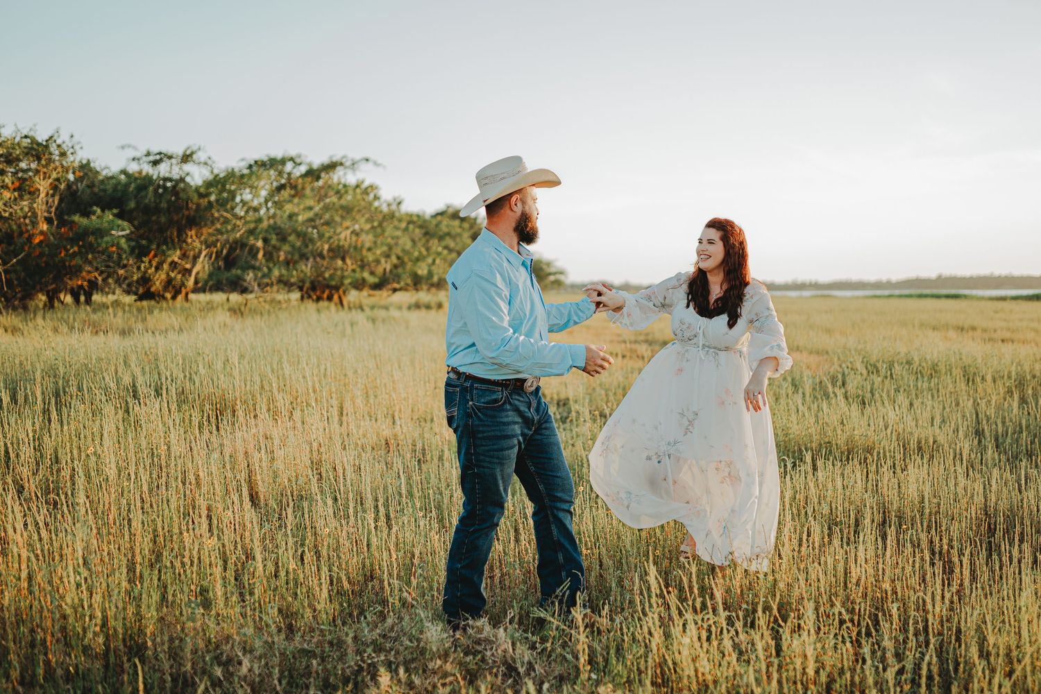 A couple in western-inspired attire dancing together in a sunlit grass meadow during golden hour.