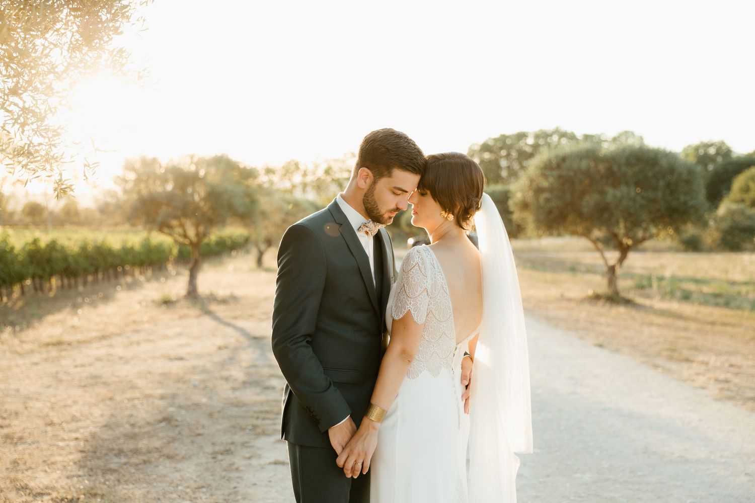 A bride and groom share an intimate moment at sunset on a dirt road near a vineyard.