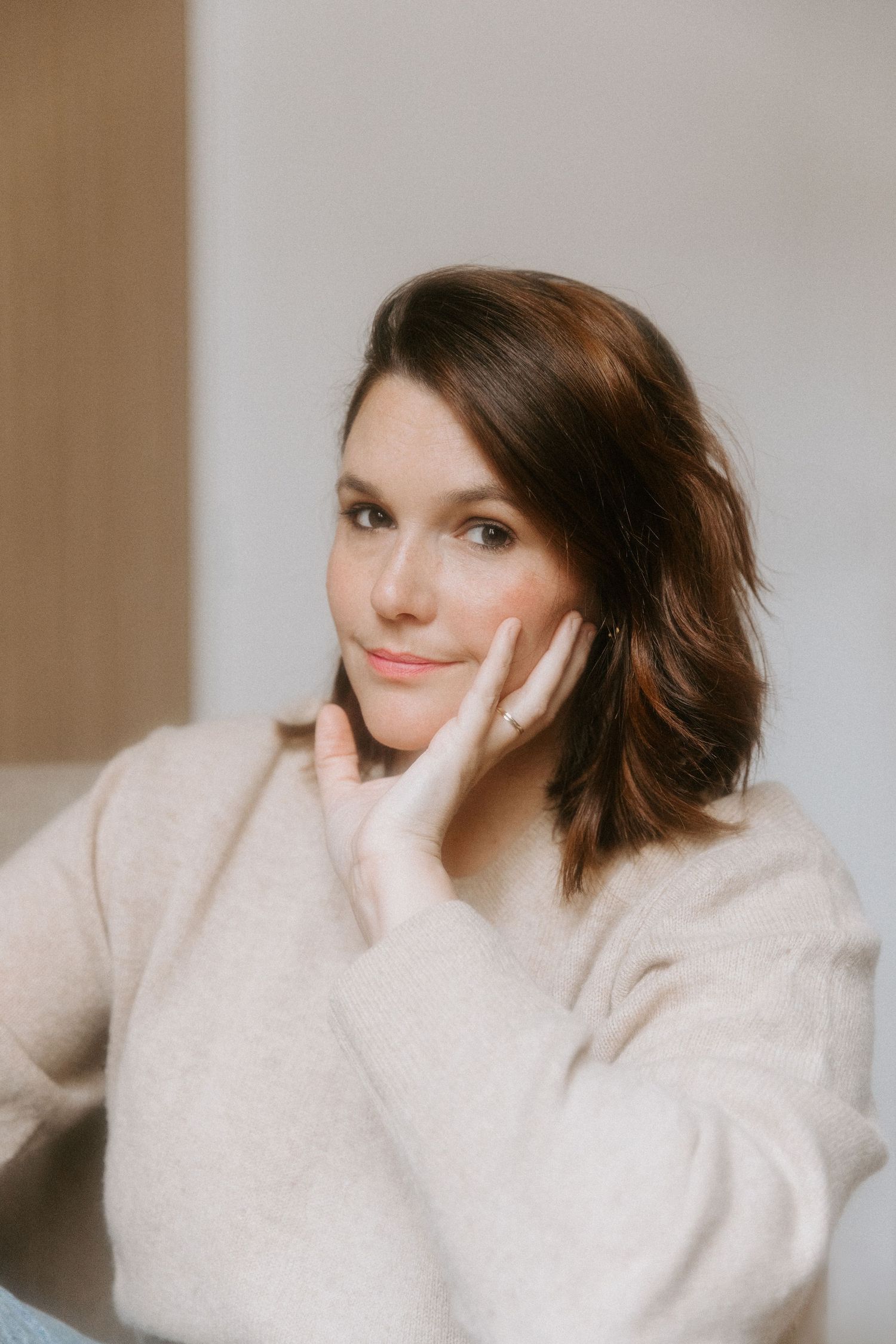 A person with shoulder-length brown hair wearing a cozy beige sweater rests their hand on their cheek while sitting indoors.