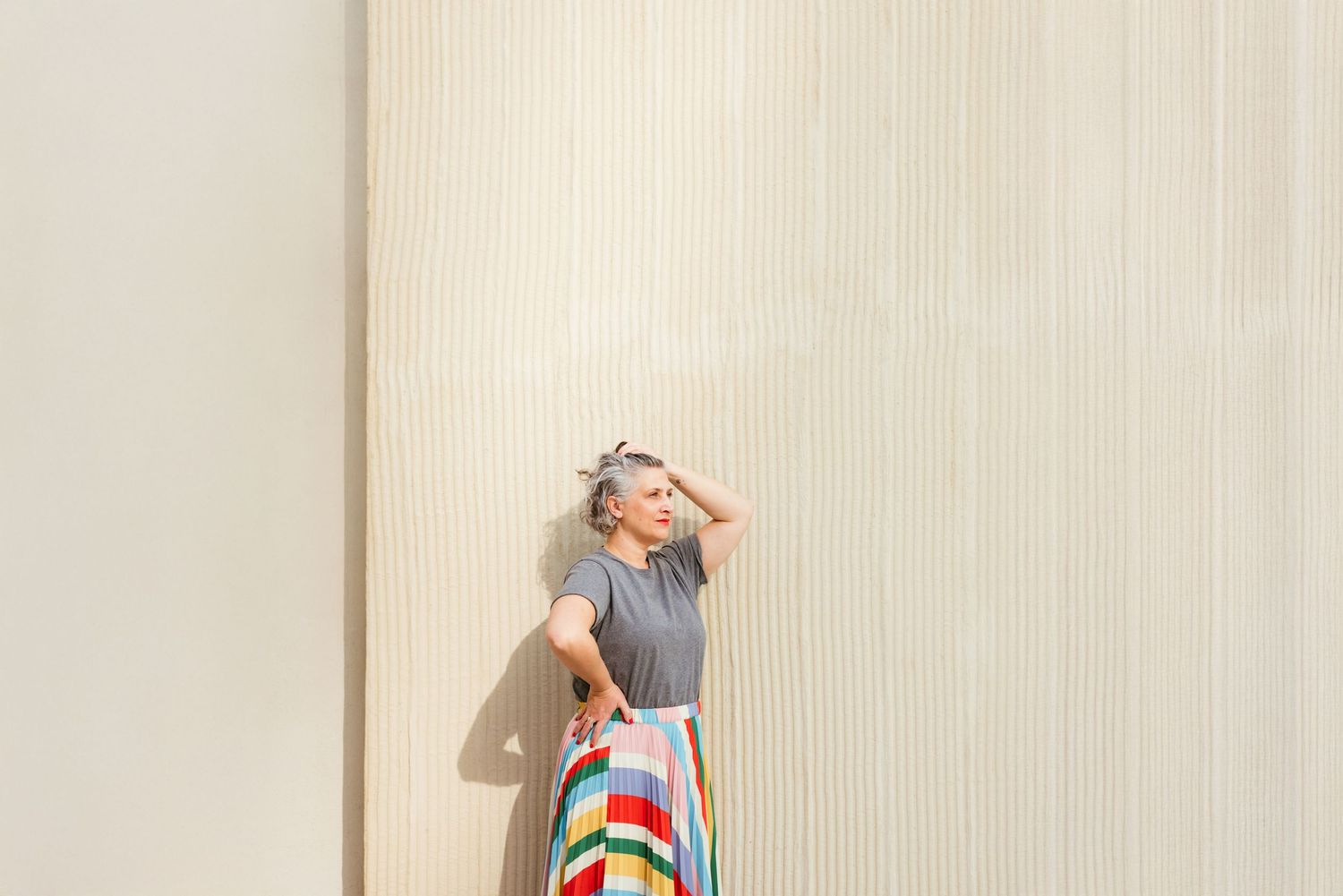 A person in a sleeveless top and colorful striped skirt stands against a beige wall casting a shadow.