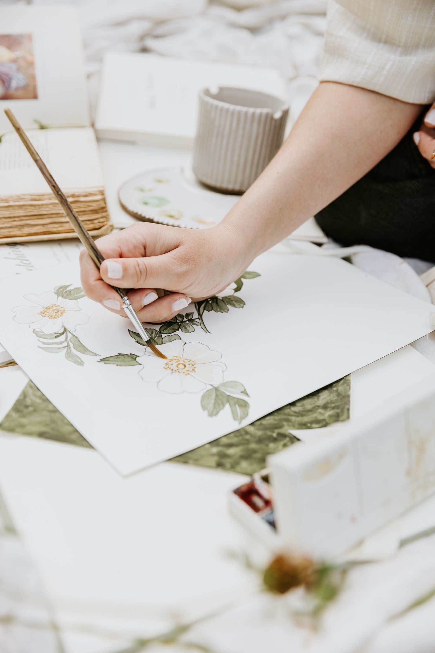 Hand holding paintbrush delicately paints botanical illustration on white paper with ceramic mug in background.