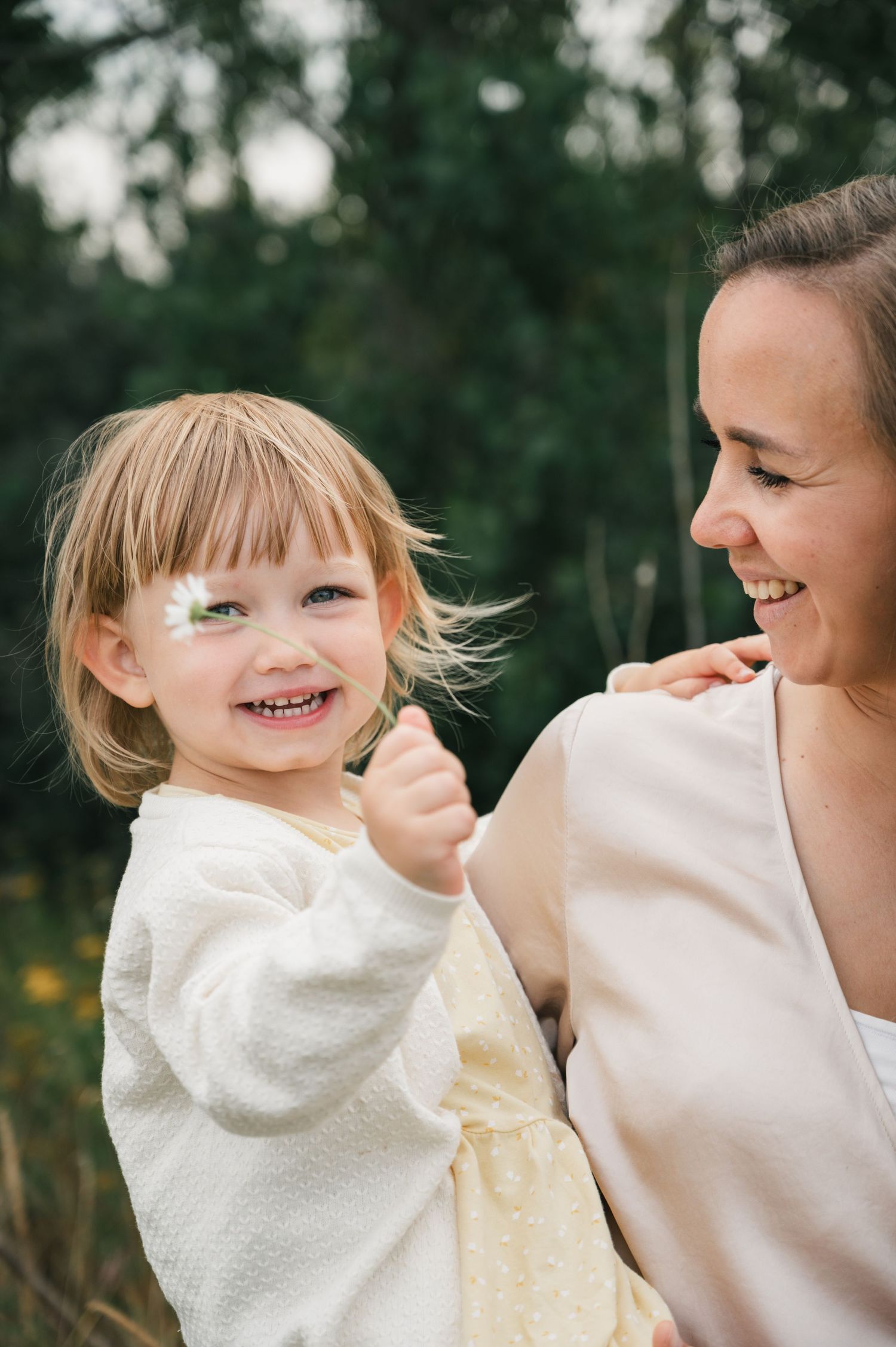 A smiling toddler girl with a daisy on her cheek being held by a laughing woman outdoors.
