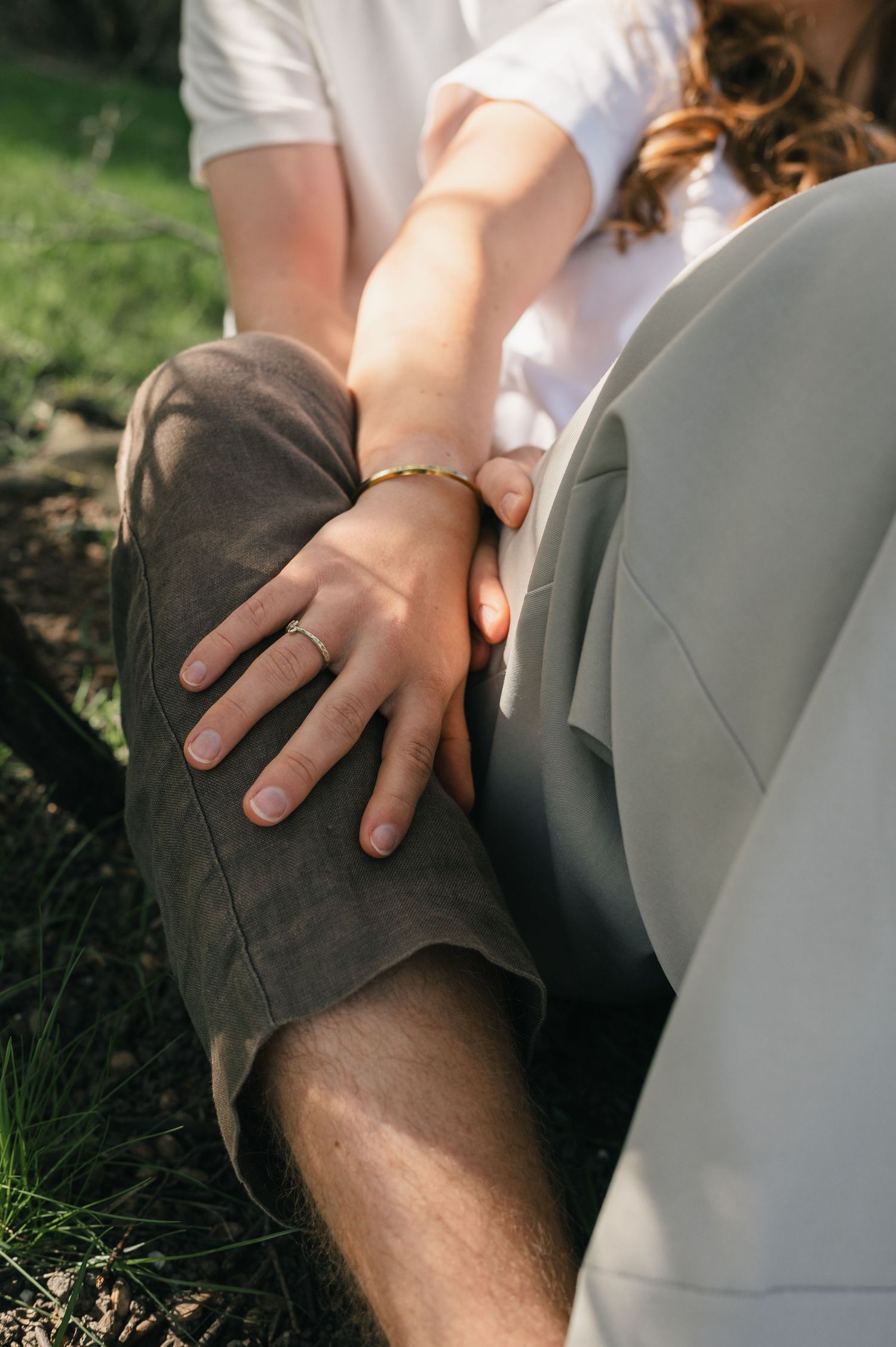 Close-up of a couple sitting outdoors, woman's hand with ring and gold bracelet resting on man's knee.