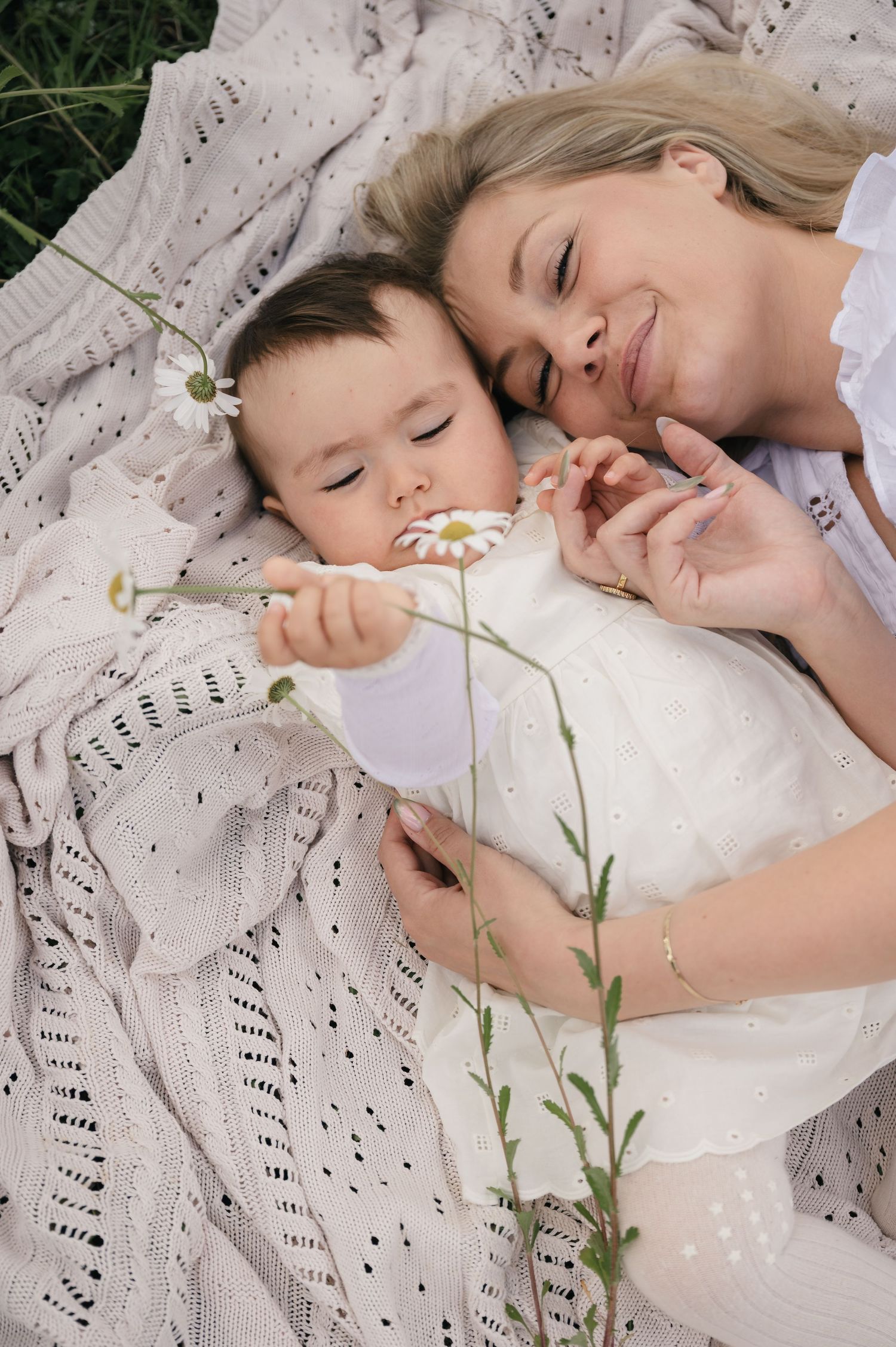 A smiling mother and baby lie on a knit blanket outdoors, sharing a tender moment with a daisy flower.