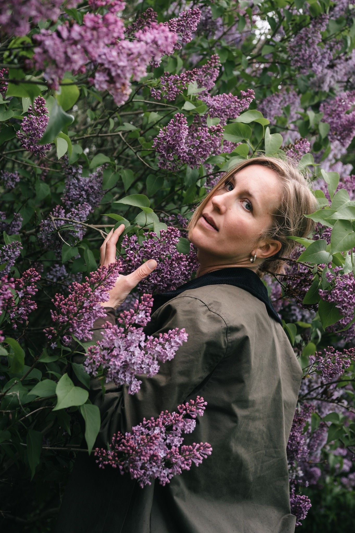 A blonde woman in an olive jacket surrounded by blooming purple lilac bushes, gazing upward in a lush garden.