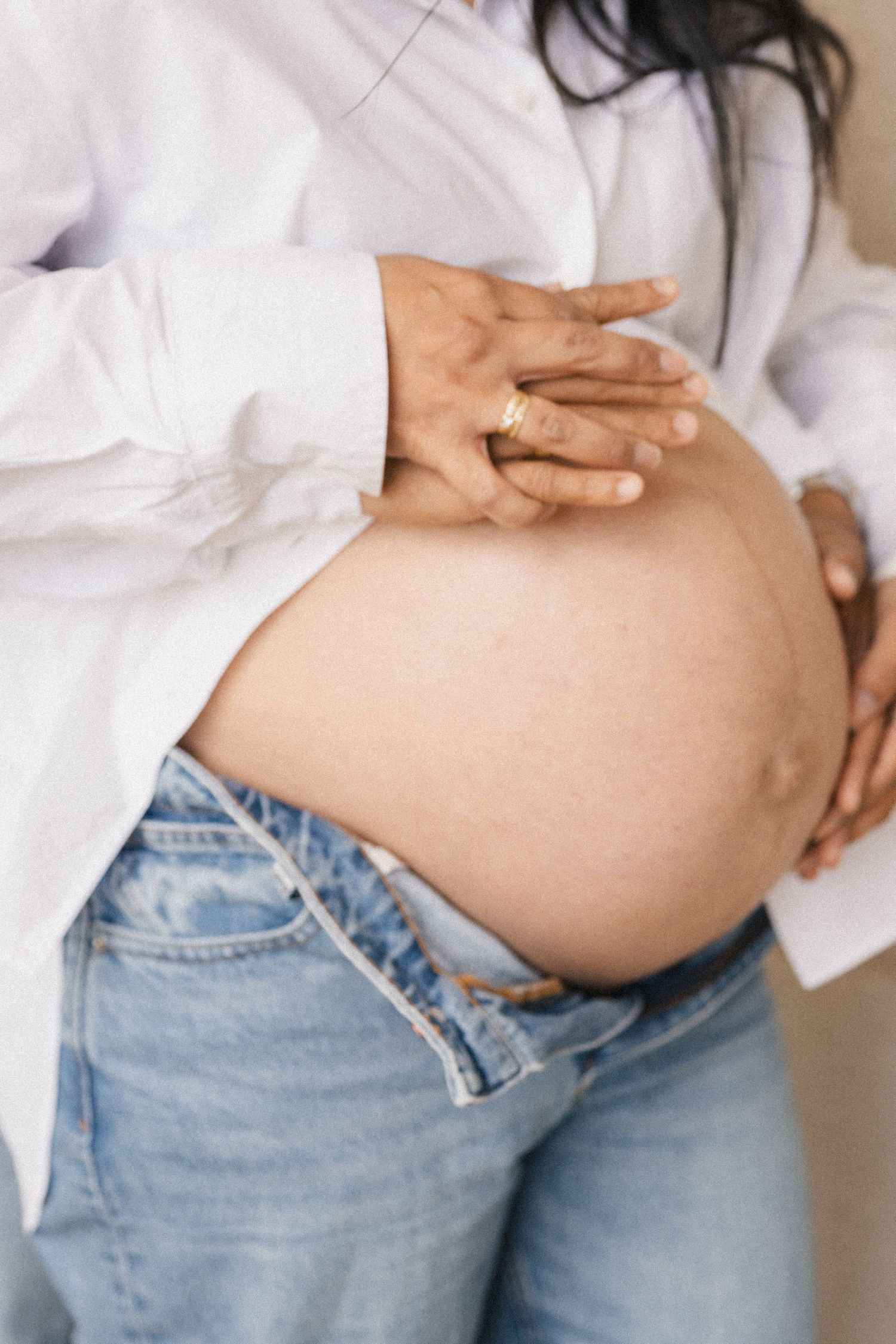 Pregnant woman in white shirt and jeans cradling her bare baby bump with both hands, wearing a gold ring.