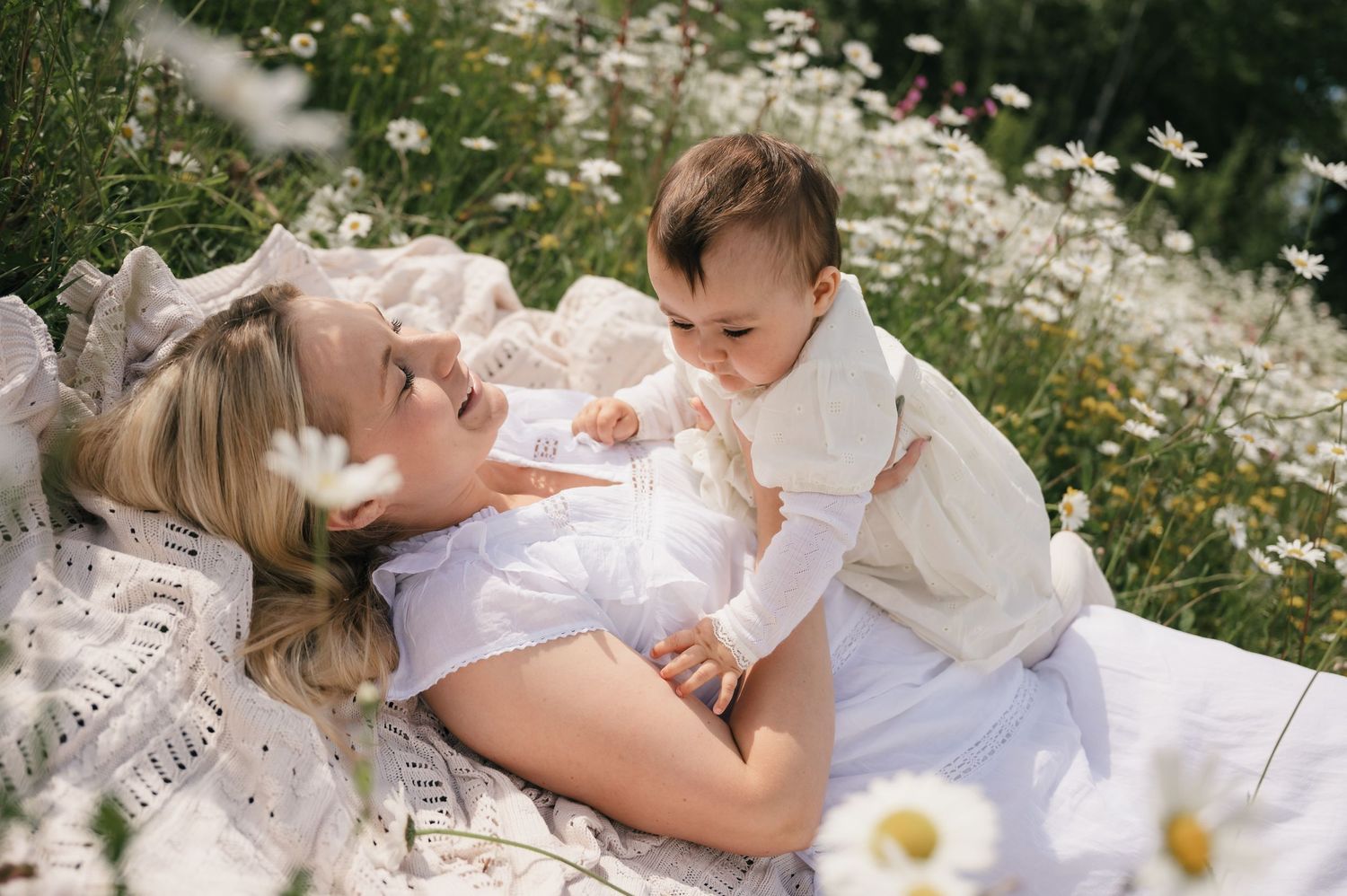 Mother lying in a daisy field, smiling at her baby sitting up in a white outfit on a sunny day.