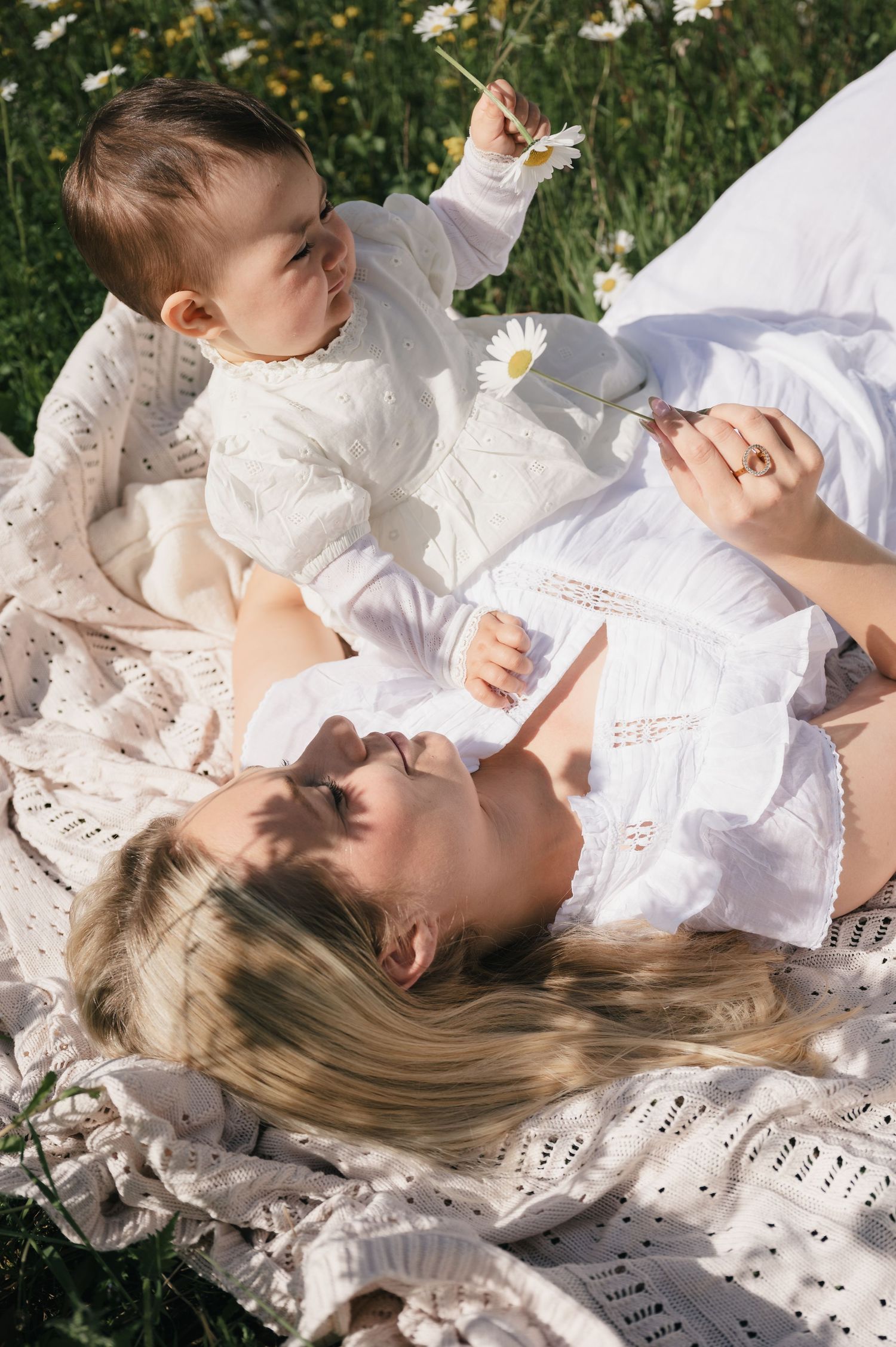Mother and baby in white outfits lying on a blanket in a flower field, sharing a daisy on a sunny day.