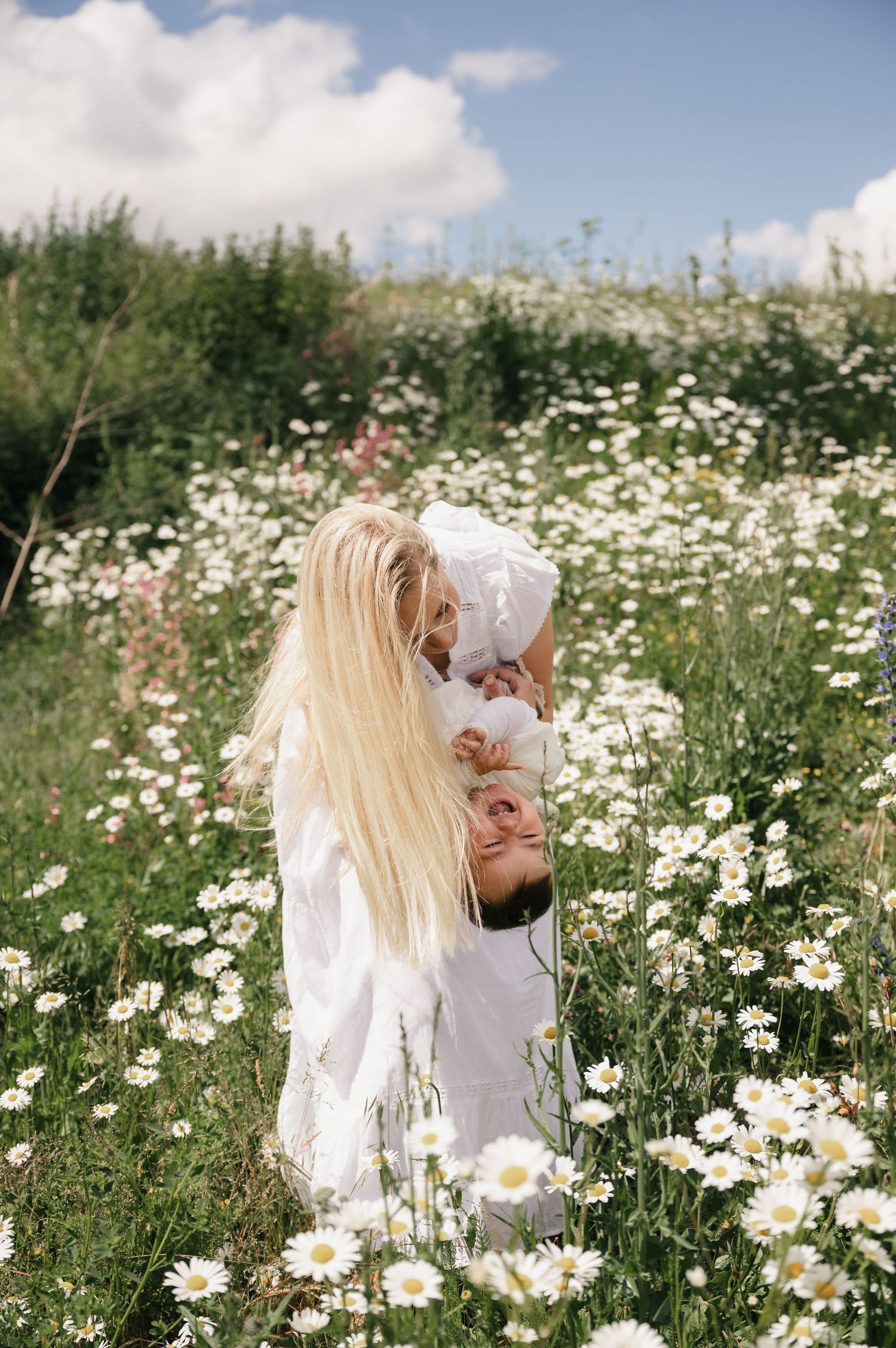 Blonde woman in white dress holding a baby in a sunny daisy field with blue sky.
