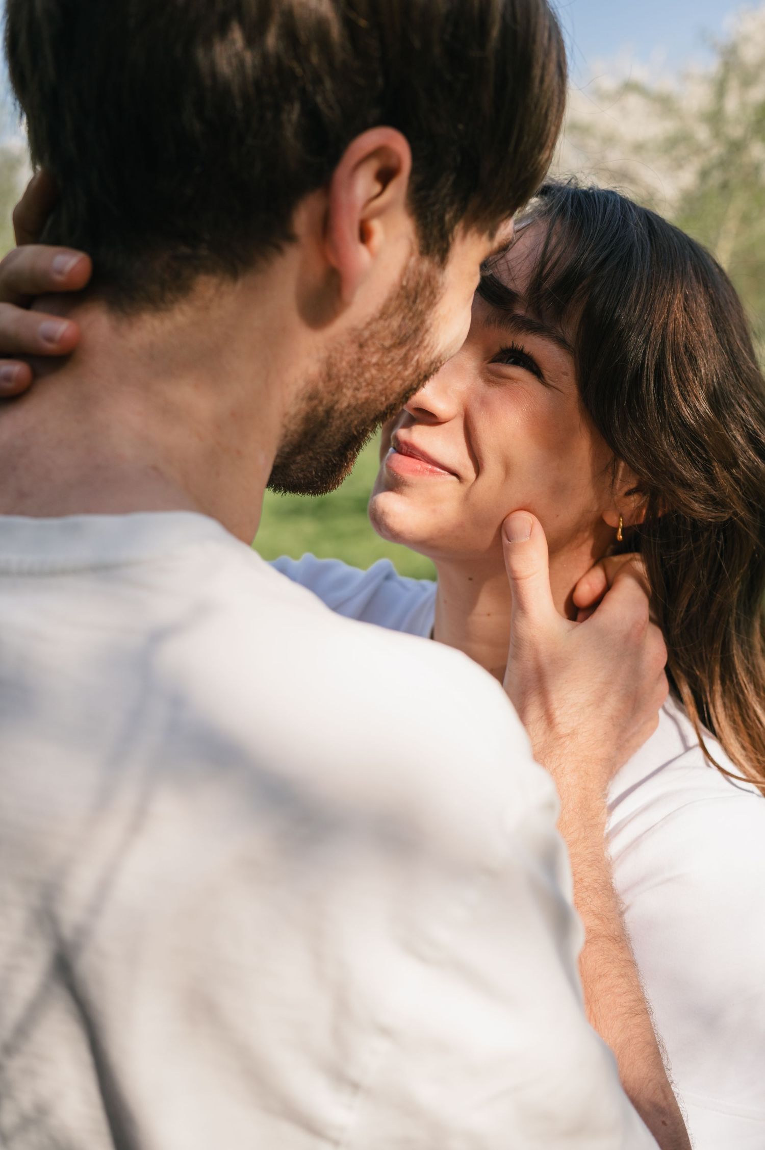 A smiling brunette woman gazes up at a man holding her face tenderly as they share a romantic moment outdoors.