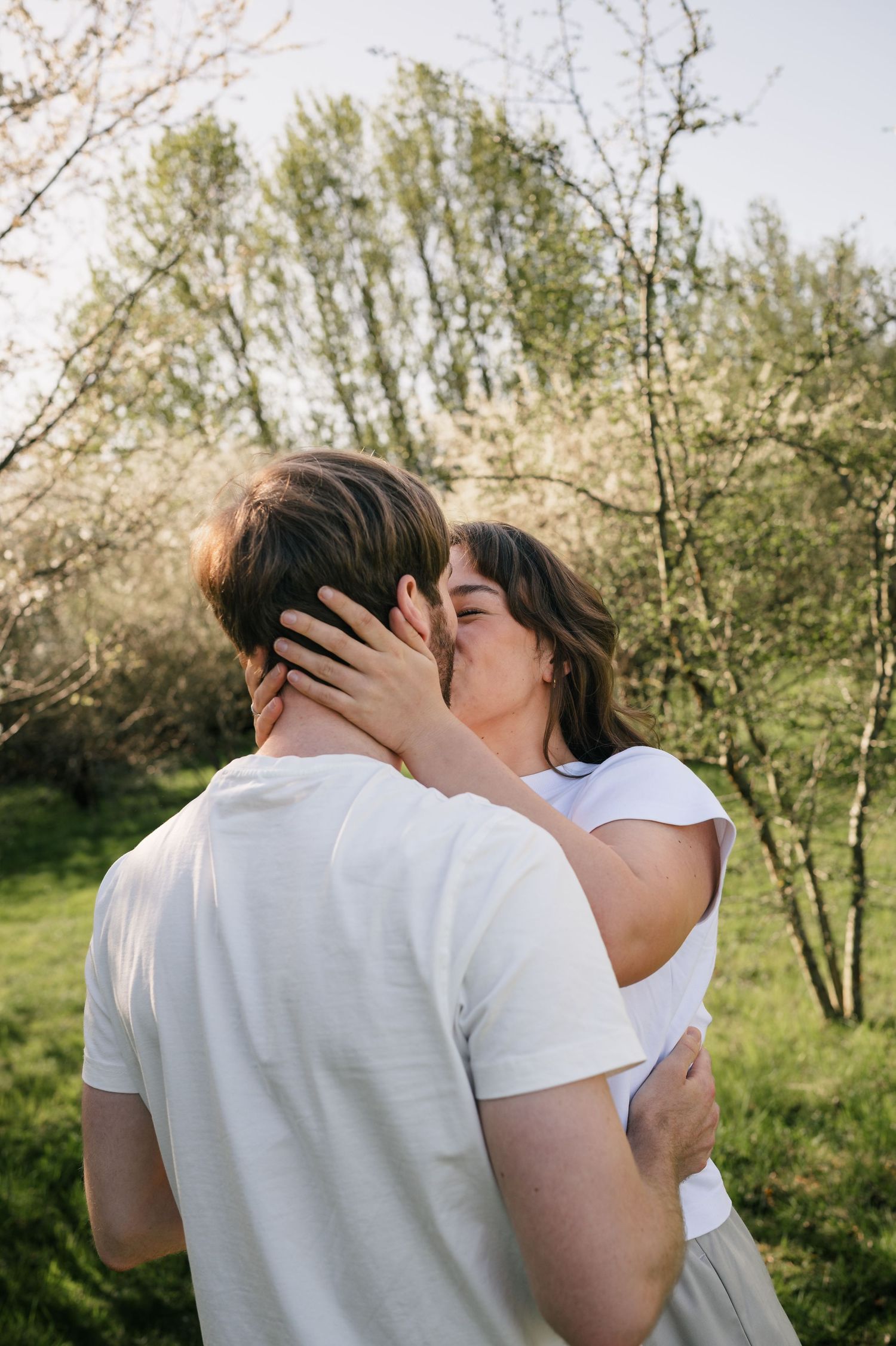 A couple sharing a romantic kiss outdoors in a green park, both wearing white shirts on a sunny day.