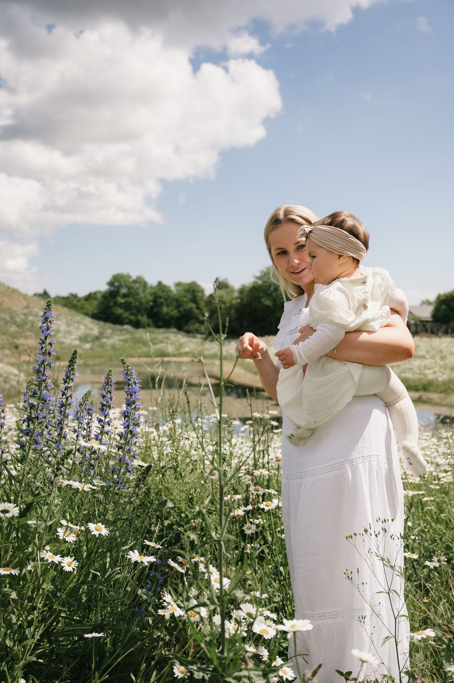 A woman in a white dress holds a baby in a flower field with lupines and daisies under a blue sky.