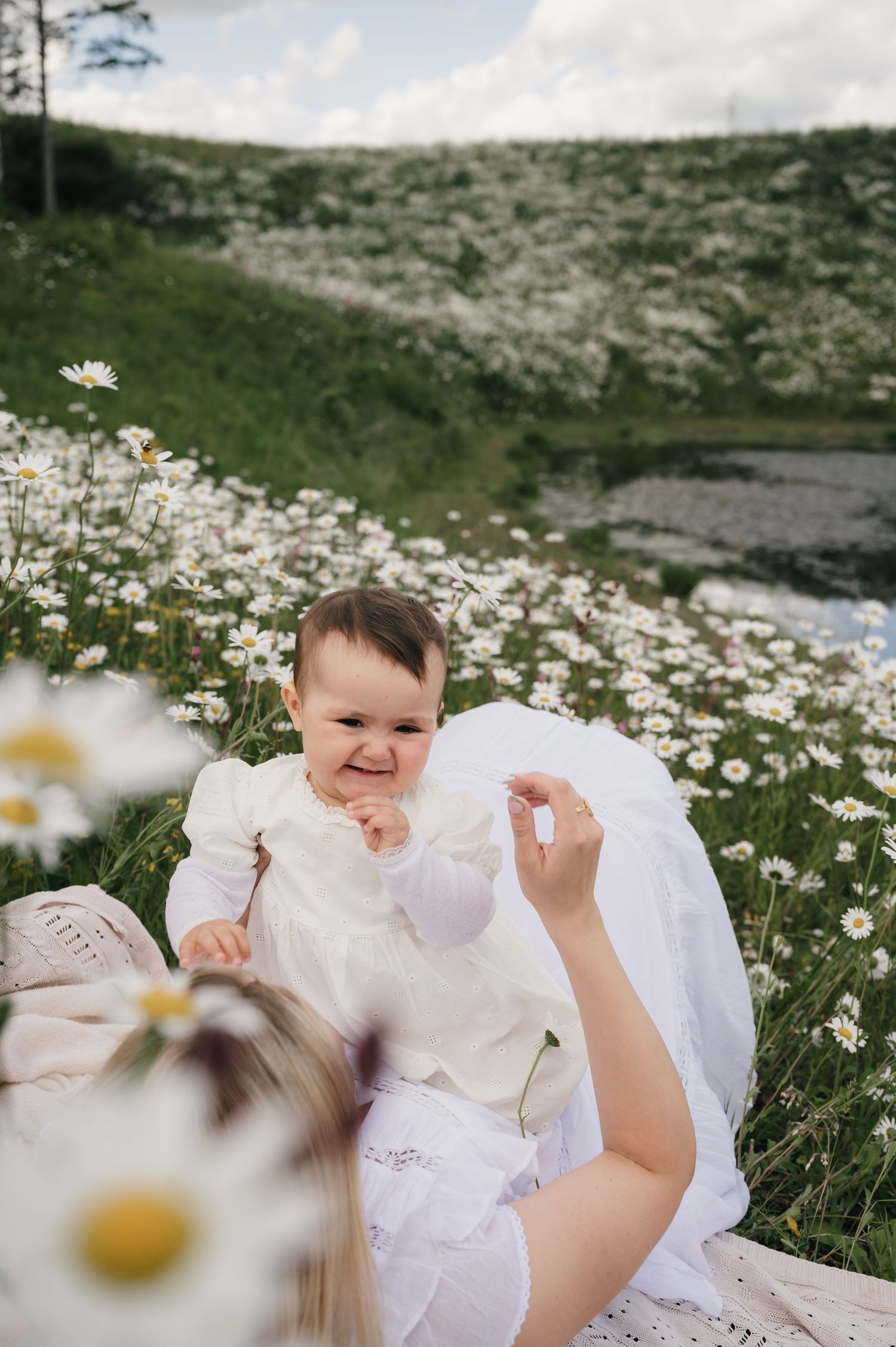 A happy baby in white outfit sits with mother in a blooming daisy field on a sunny day.
