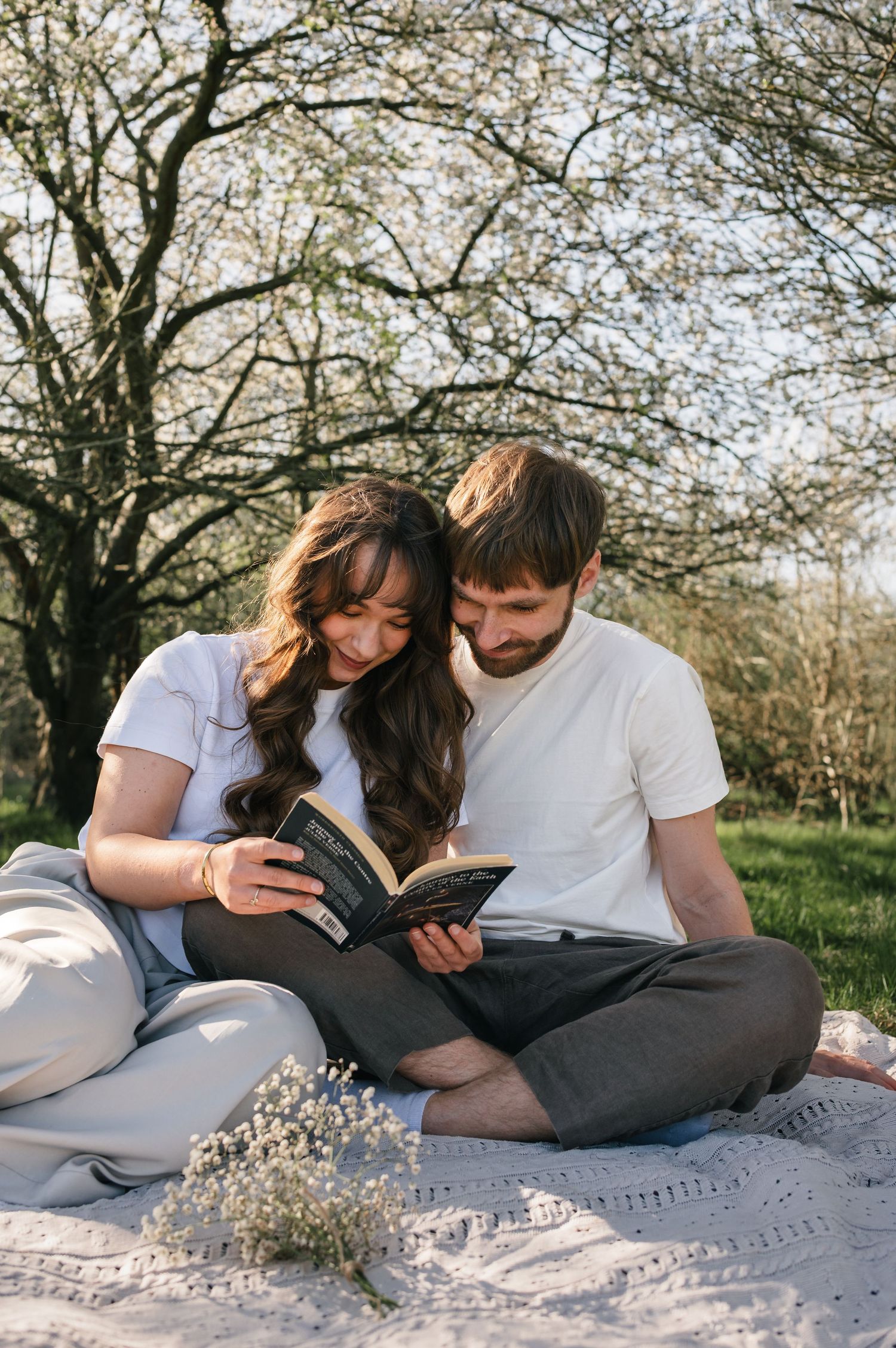 Couple sitting on a blanket outdoors reading a book together near blooming trees on a sunny day.