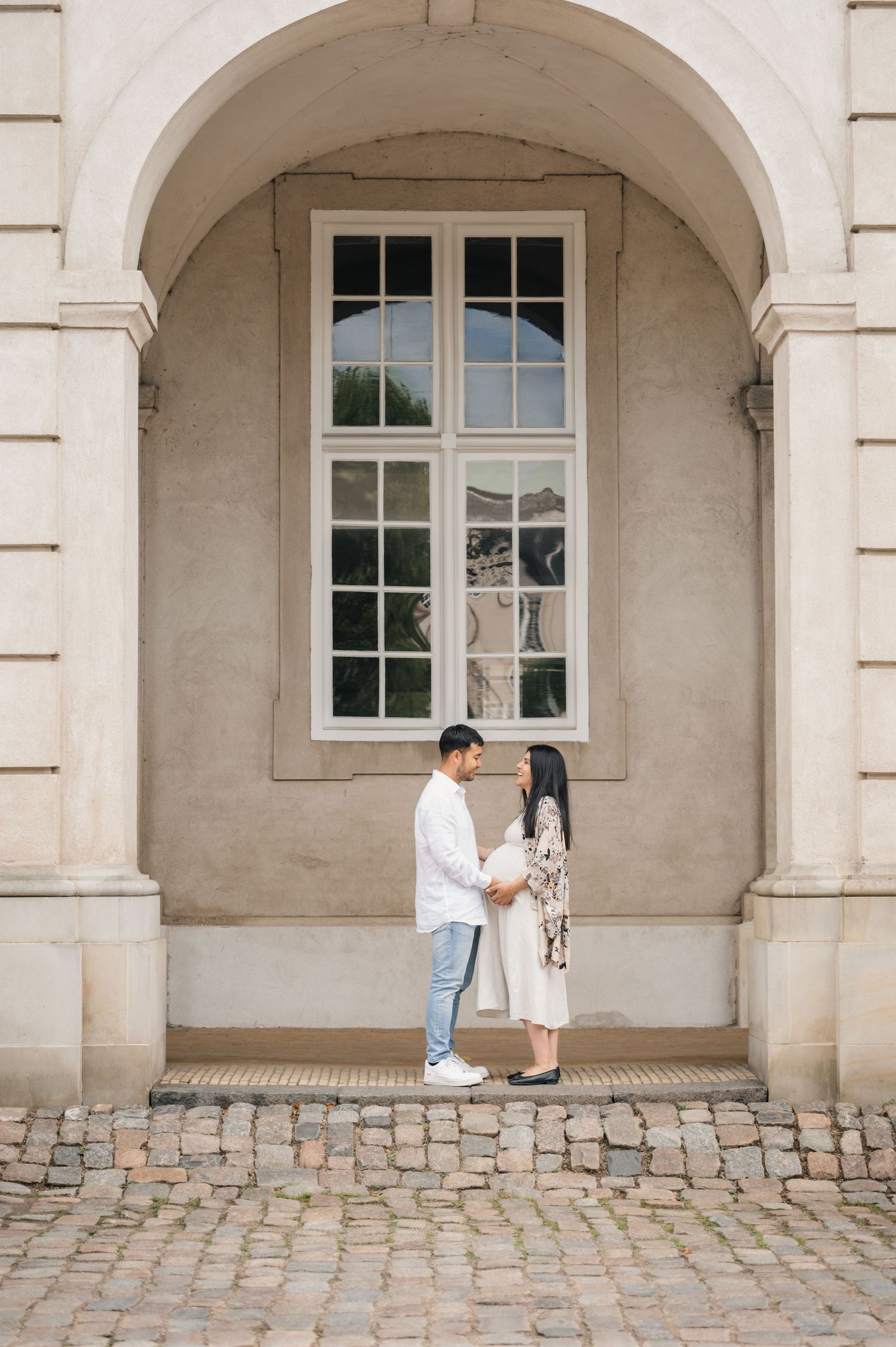 Couple holding hands facing each other under a stone arch on cobblestone, elegant building backdrop.