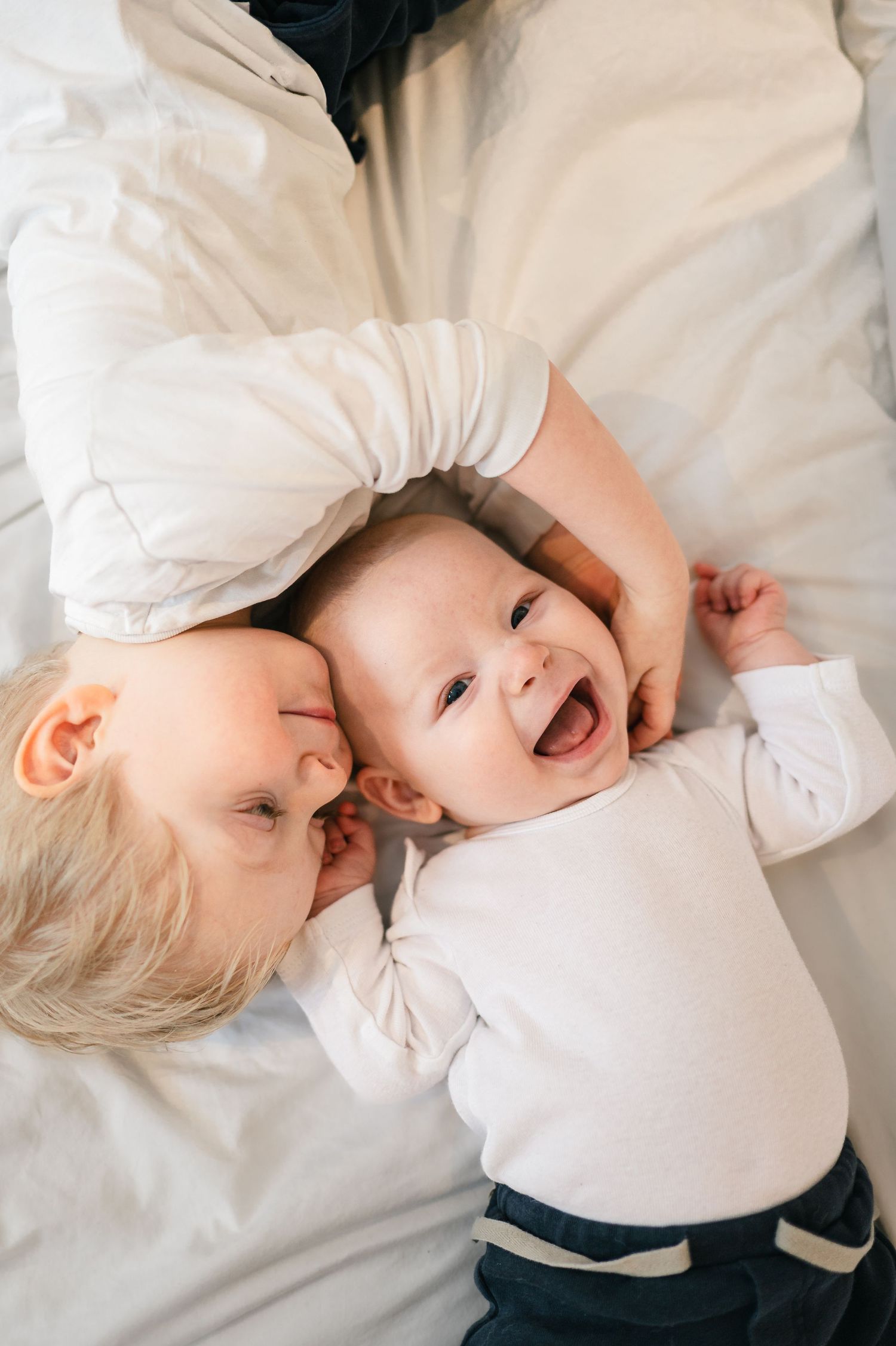 En baby og hans storebror, begge klædt i hvidt og liggende på hvidt sengetøj, smiler og interagerer sødt med hinanden i et spontant øjeblik