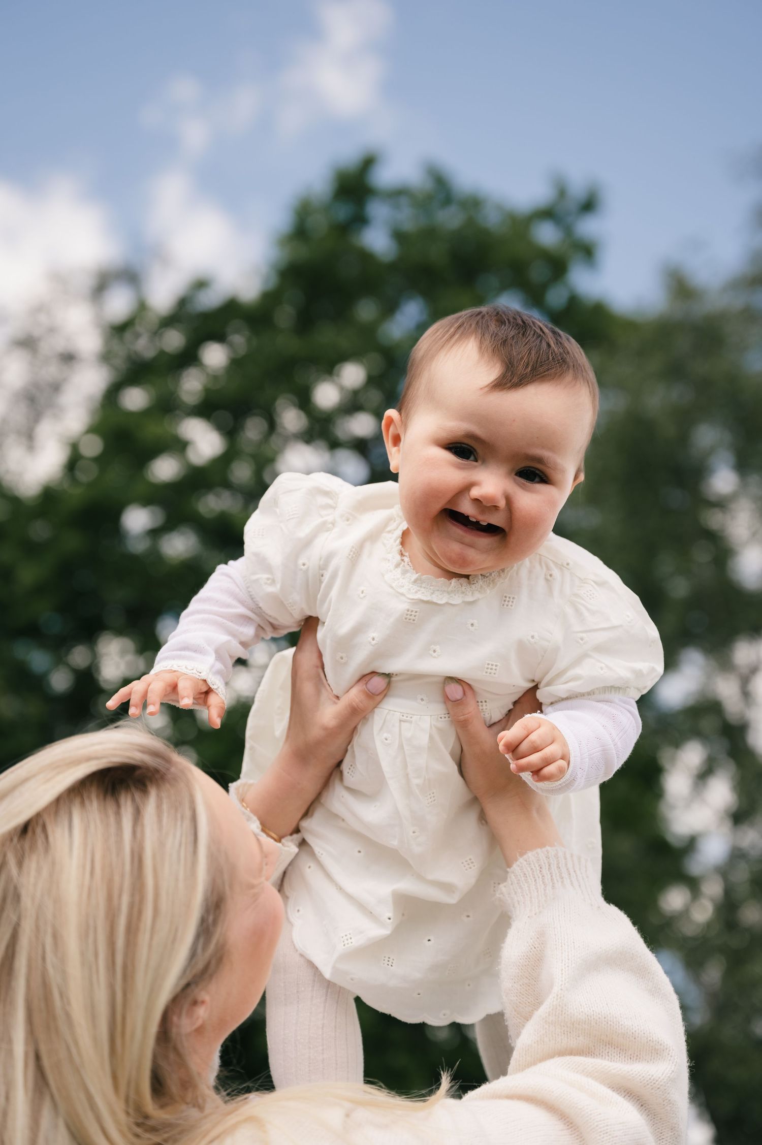 A joyful baby in a white dress is lifted up by a smiling blonde mother outdoors with green trees behind them.