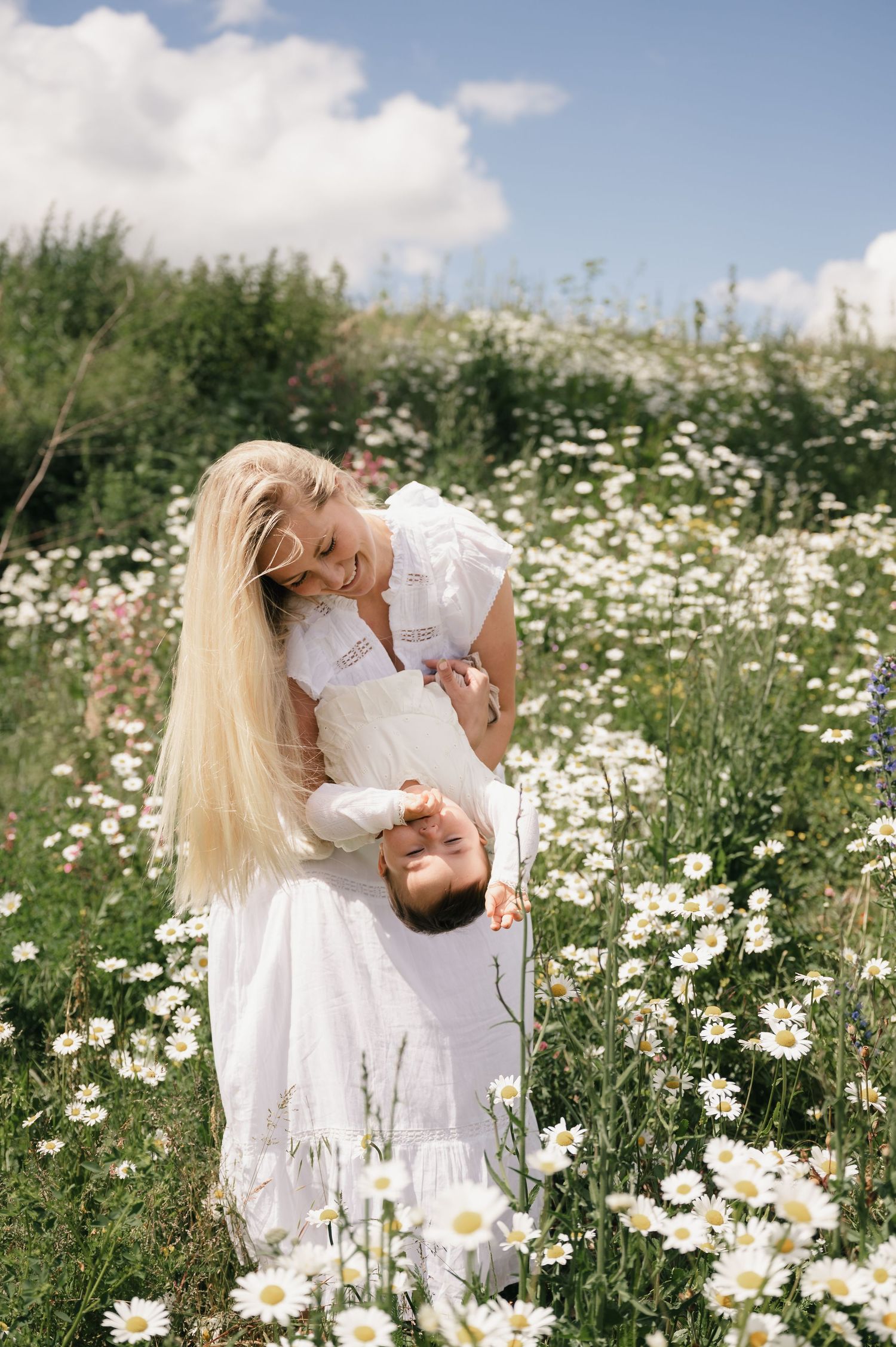 A smiling blonde woman in a white dress playfully holds a child upside down in a sunny daisy field.
