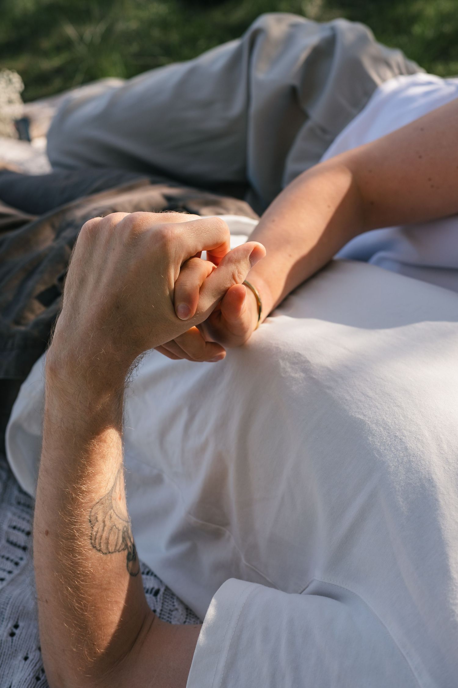 Two people holding hands tenderly outdoors, one with a tattoo on their wrist, lying on a white blanket.