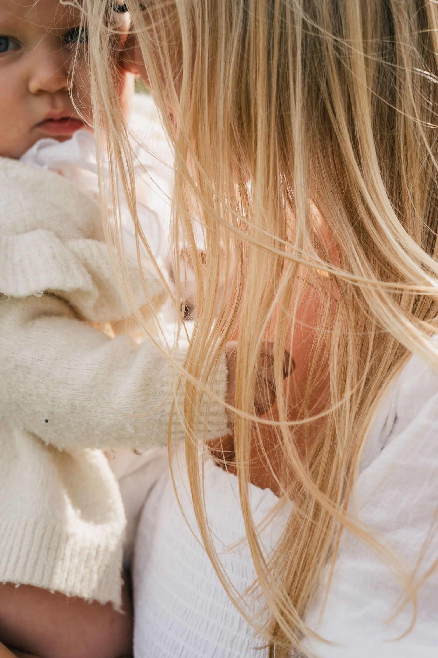 A blonde woman with long hair gently holds a young toddler dressed in a white knit sweater outdoors.